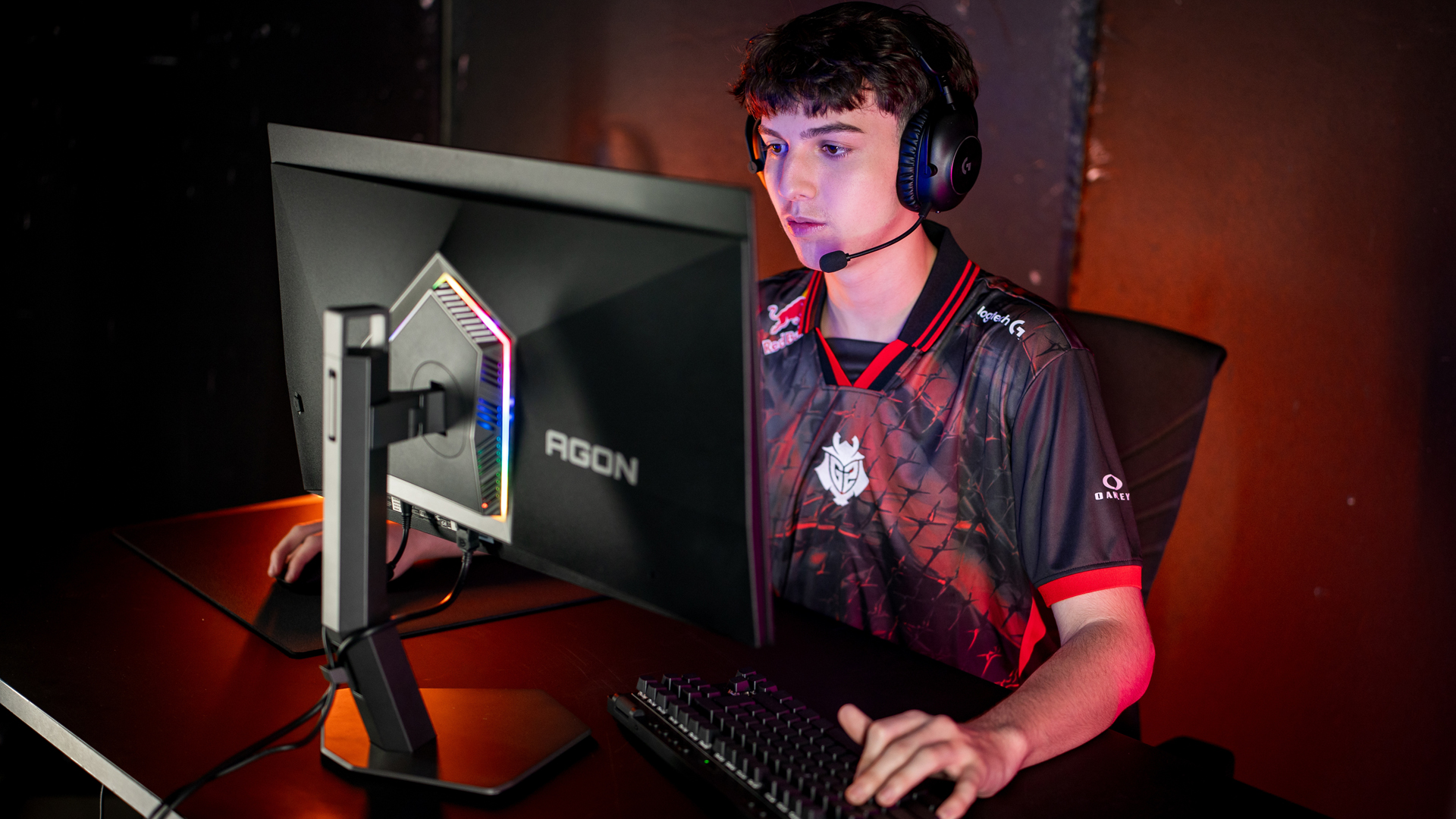 Person gaming on an AGON monitor, wearing Logitech headphones and a jersey with Red Bull and Oakley logos, in a dimly lit room.