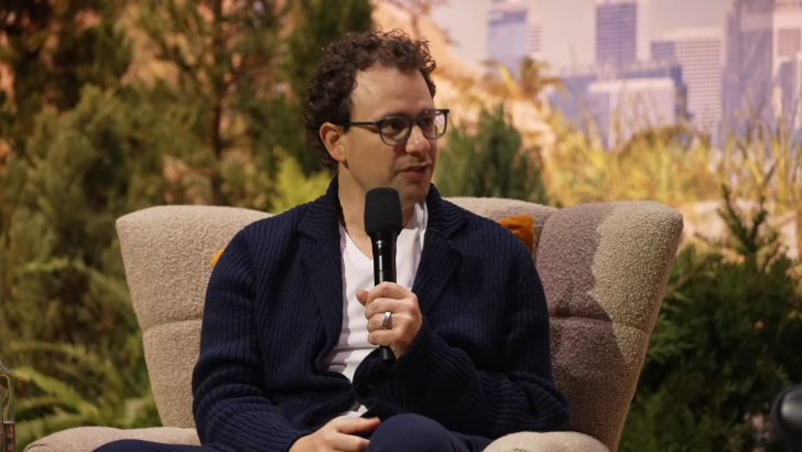 Man holding a microphone sitting on a chair with a nature backdrop.