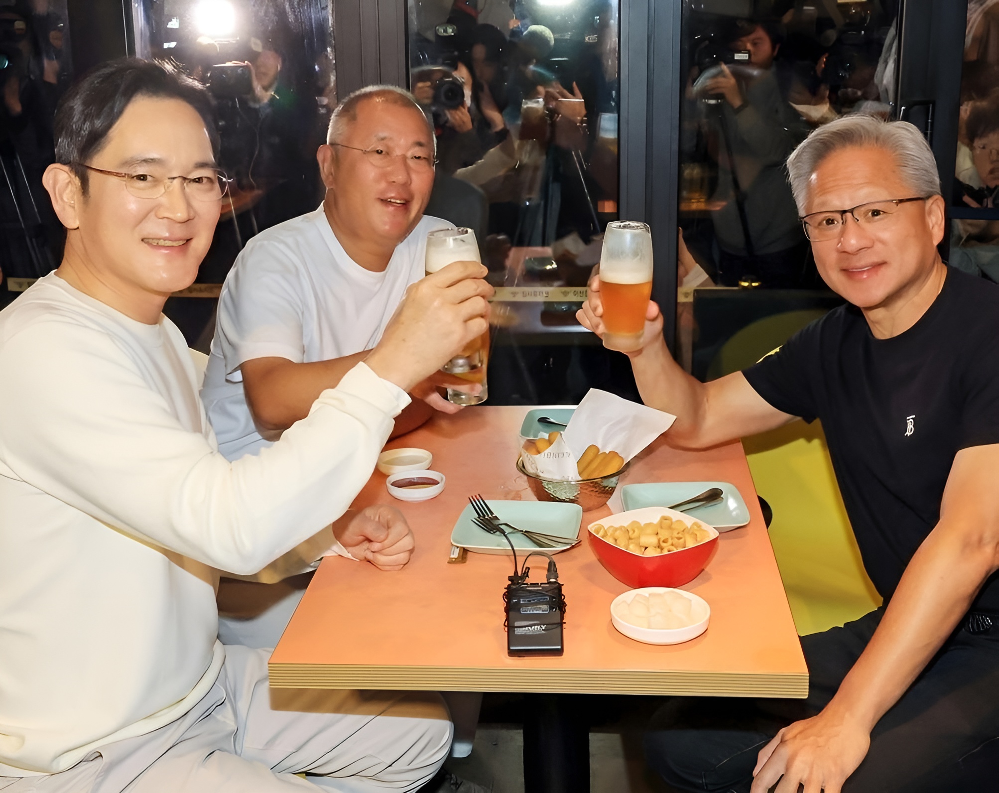 Three people raising beer glasses at a restaurant table with a Sony handheld recorder visible.