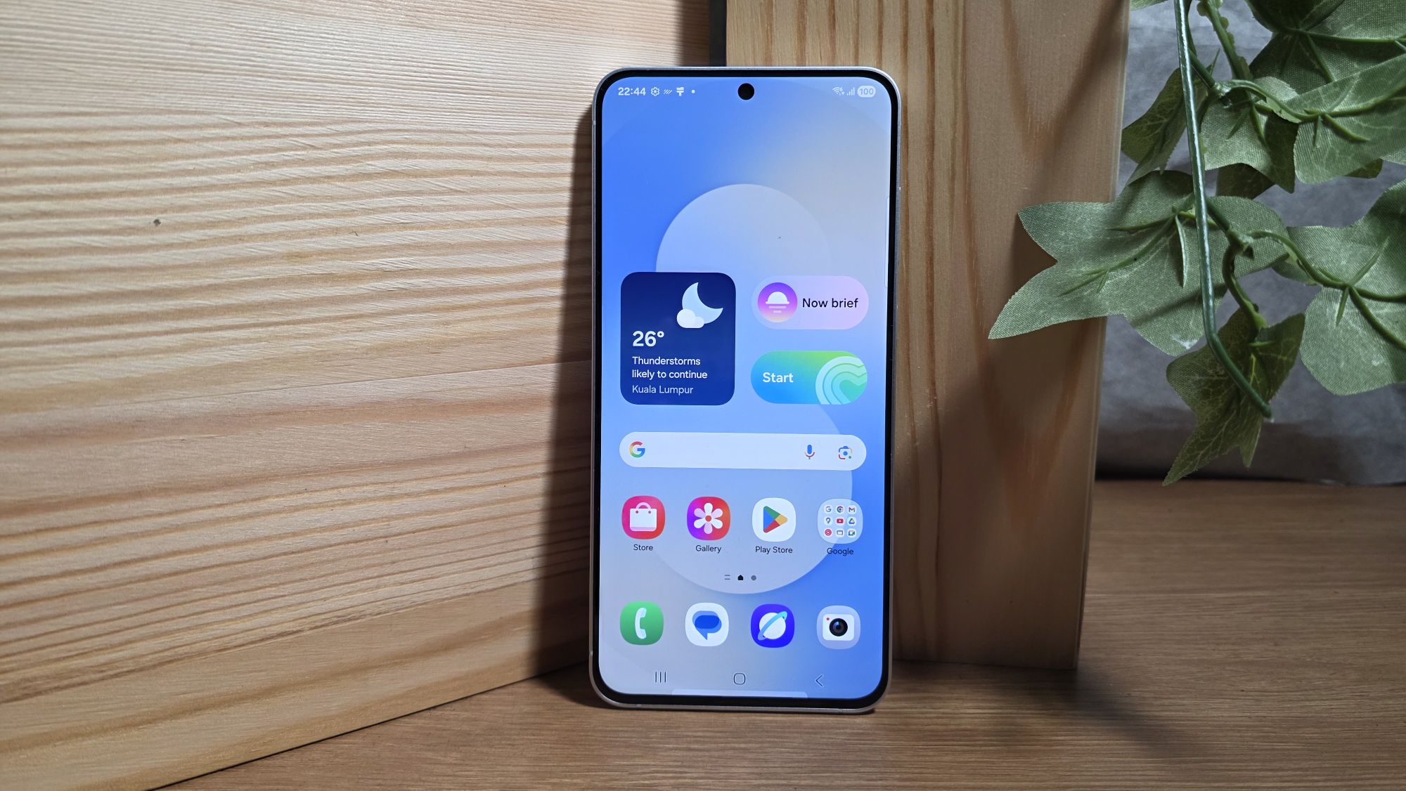 Samsung smartphone displaying weather app with 26° Thunderstorms likely to continue Kuala Lumpur on a wooden table beside a plant.