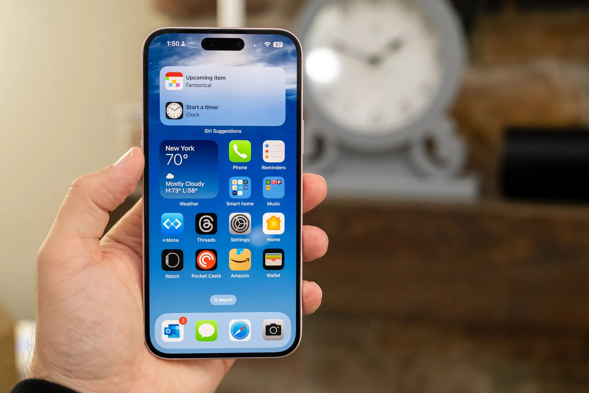 A hand holding an iPhone 15 Pro displaying a home screen with various apps, including Fantastical and Threads, and a weather widget showing 'New York 70°'.