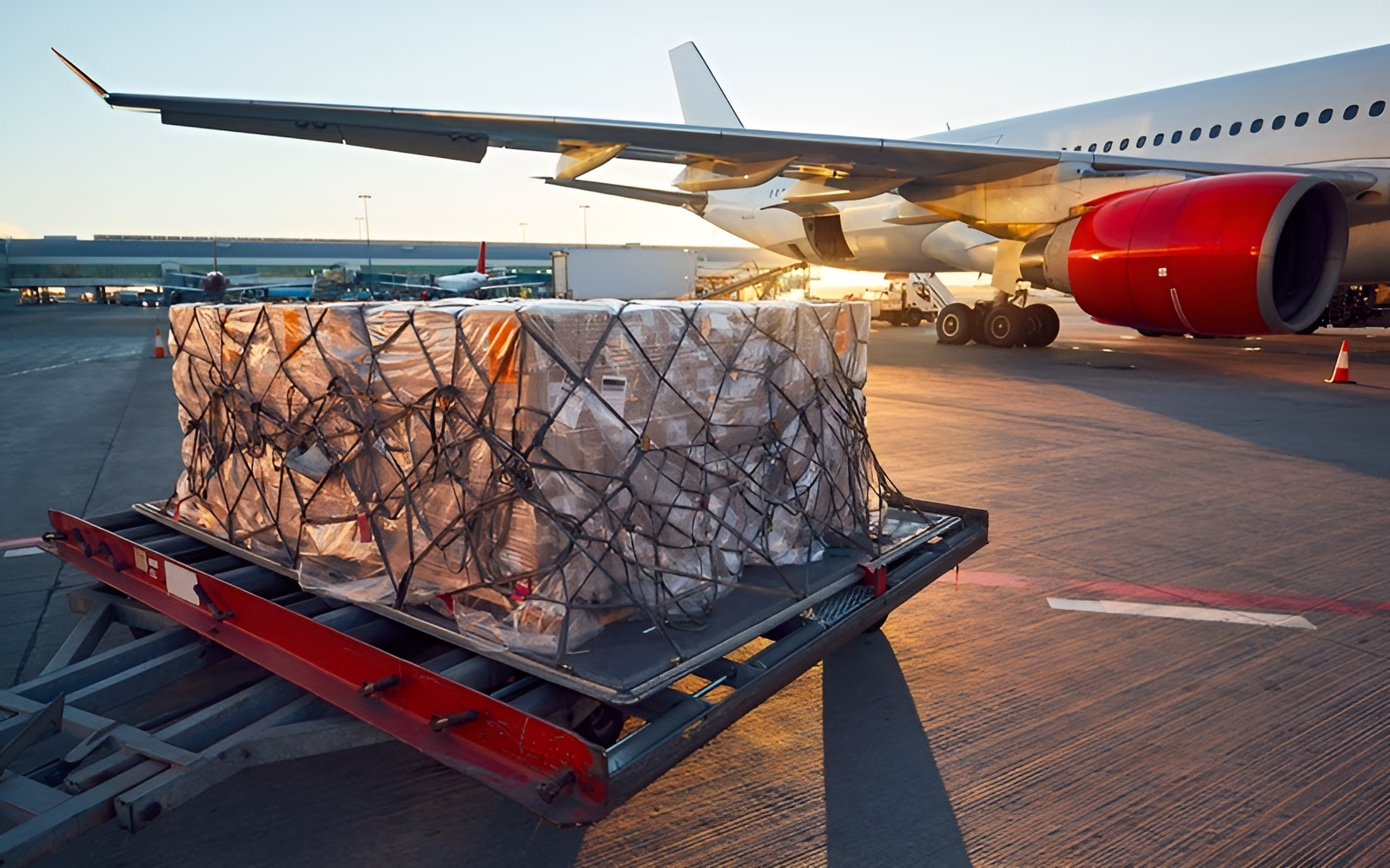 A cargo pallet is loaded onto a plane with red engine covers on an airport tarmac during sunset.
