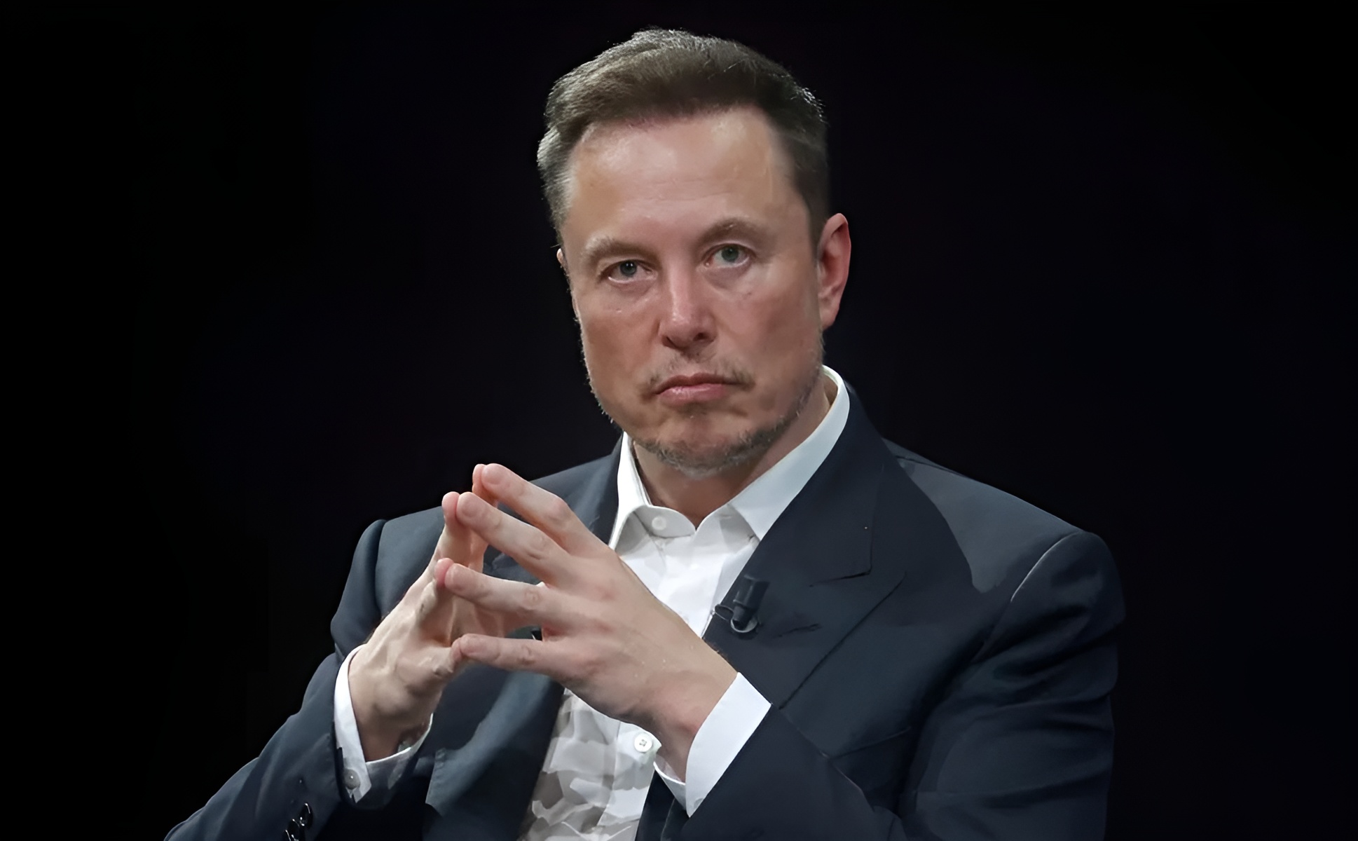 A person in a suit sits against a dark background, with fingers interlocked in a contemplative gesture.