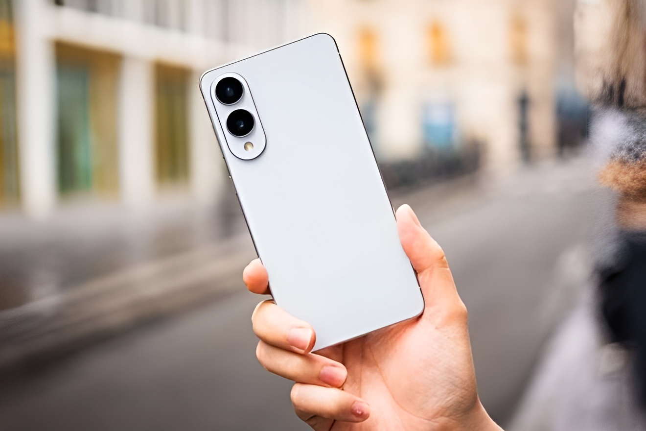 A person holds a white Samsung smartphone with a dual-camera setup outdoors.