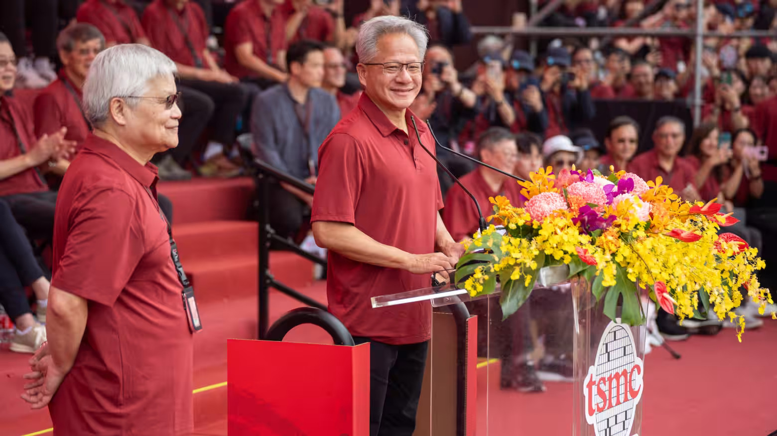 A man in a red shirt speaking at a podium with a 'tsmc' logo, surrounded by colorful flowers, in front of a group of people also wearing red shirts.
