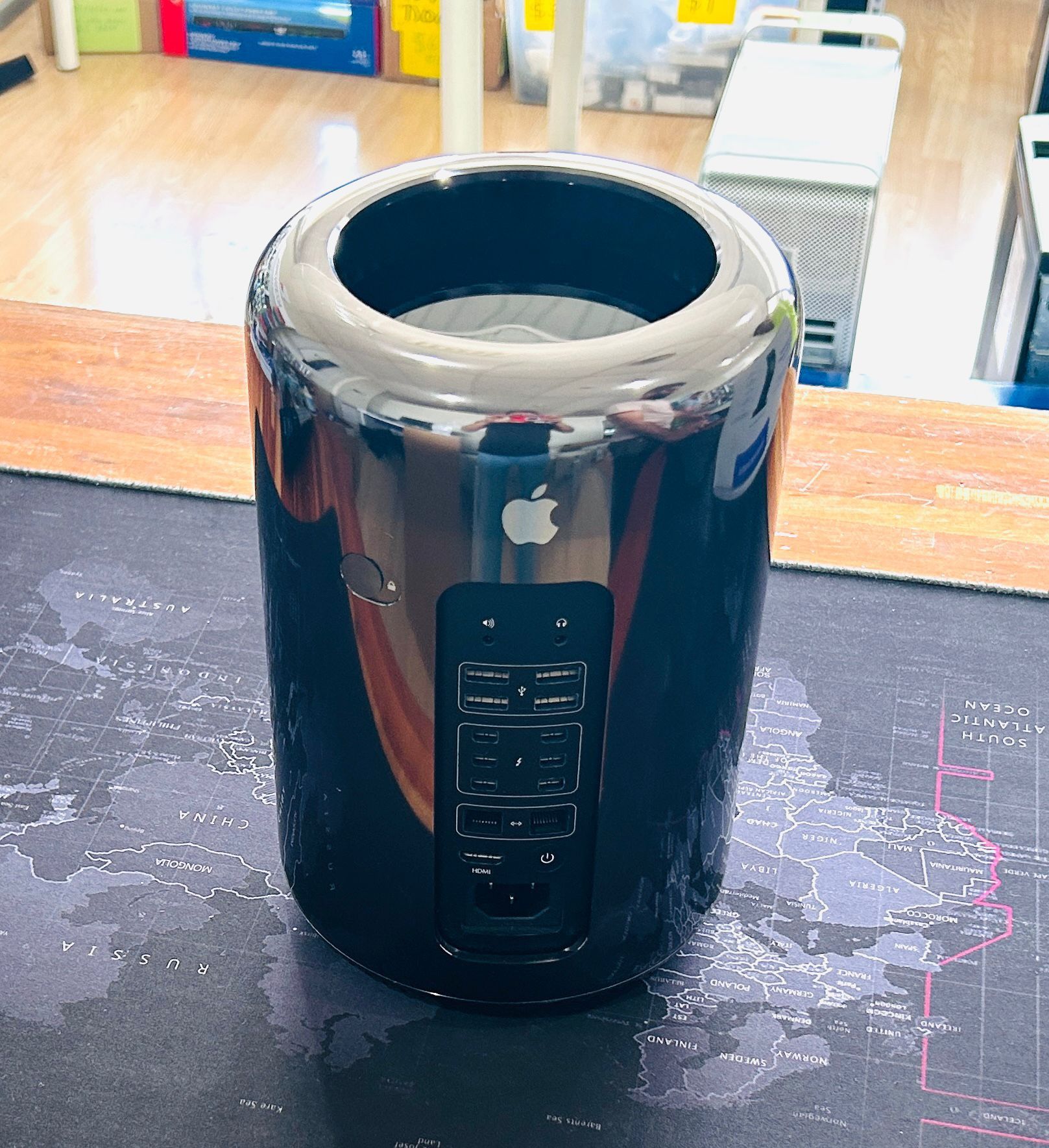 The image shows an Apple Mac Pro (2013) with its ports visible on a desk featuring a world map design.