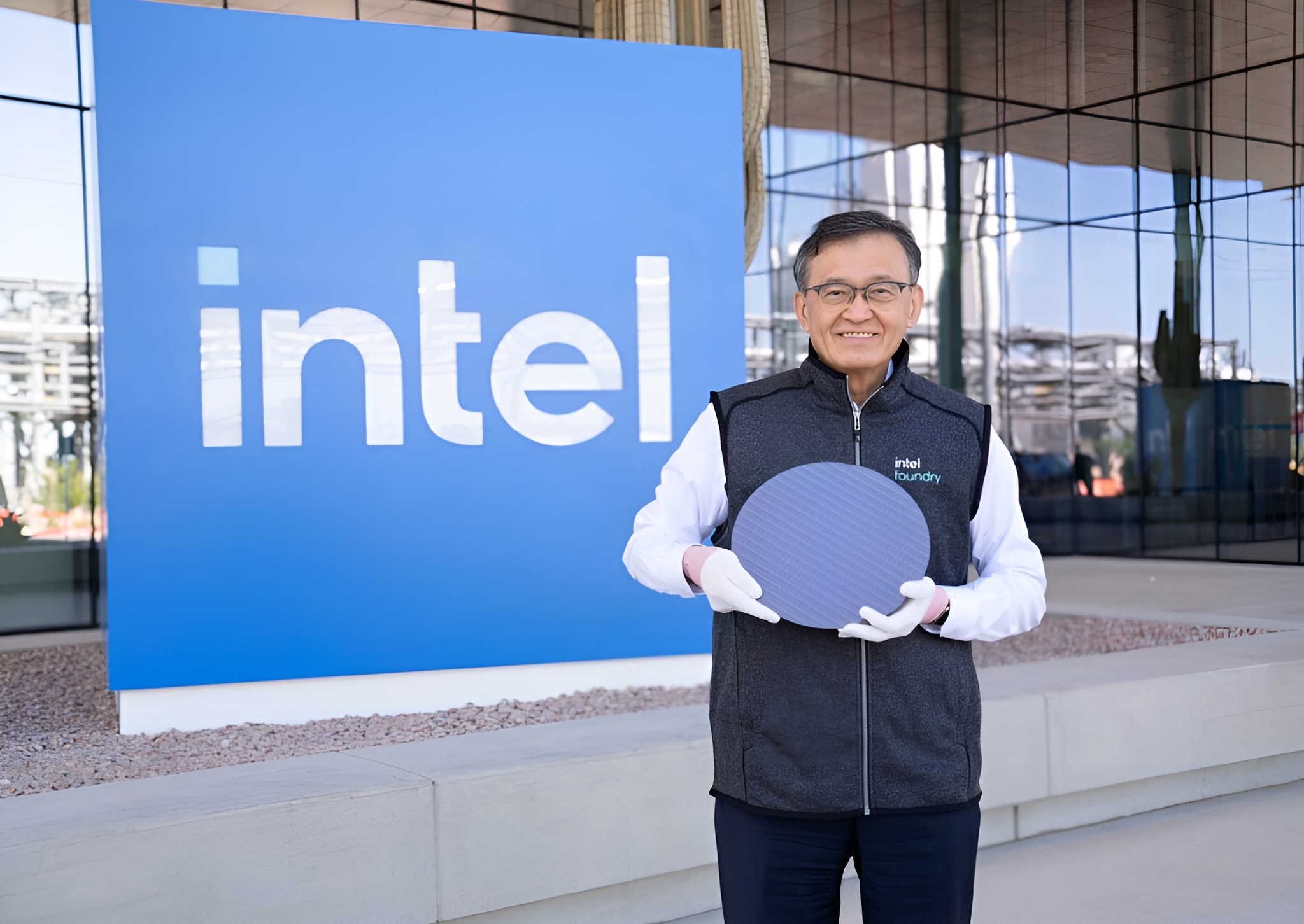 A person standing in front of an Intel sign, holding a semiconductor wafer, wearing an 'intel foundry' vest.