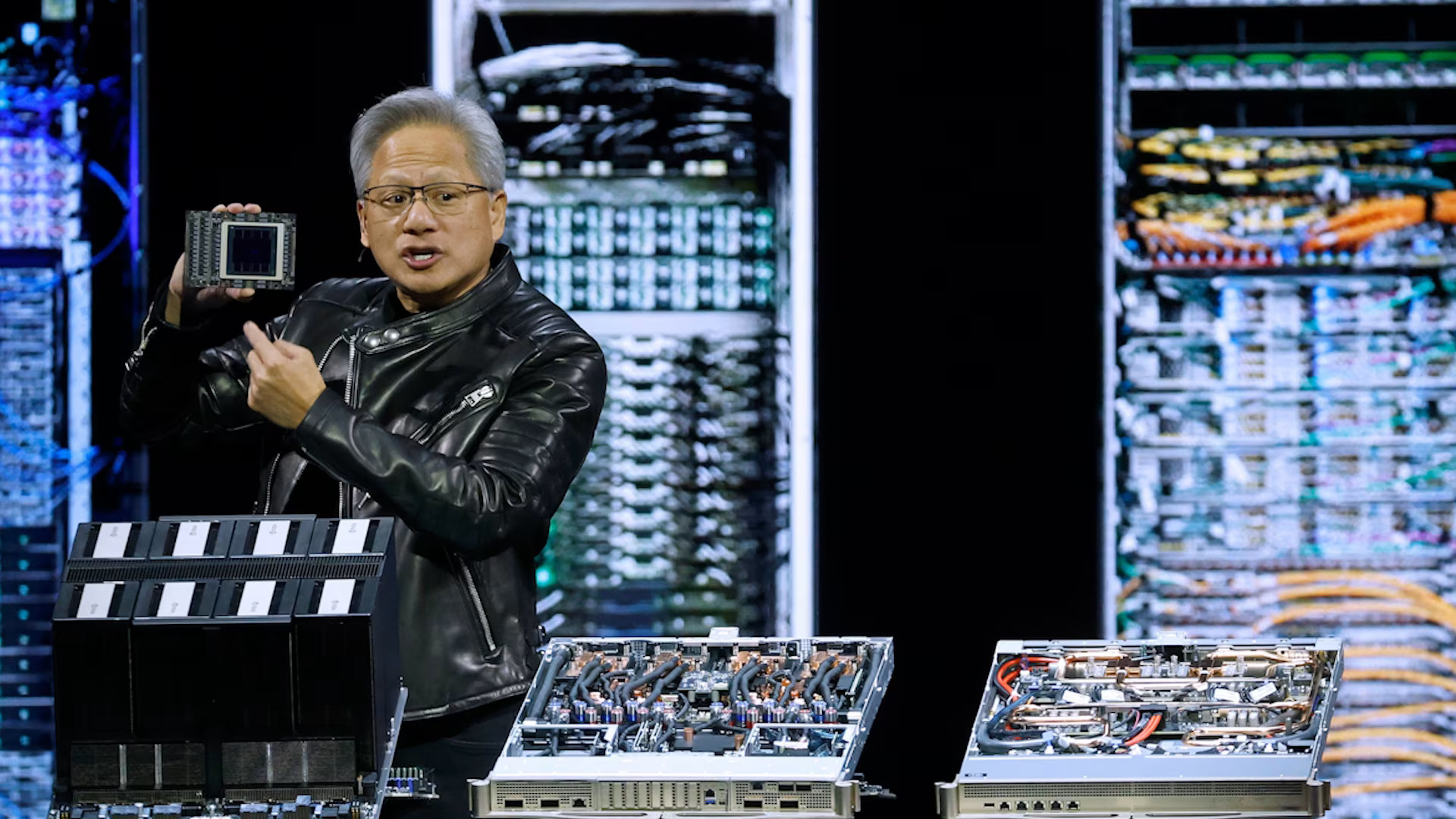 Man showcasing NVIDIA GPU on stage with server racks in the background.