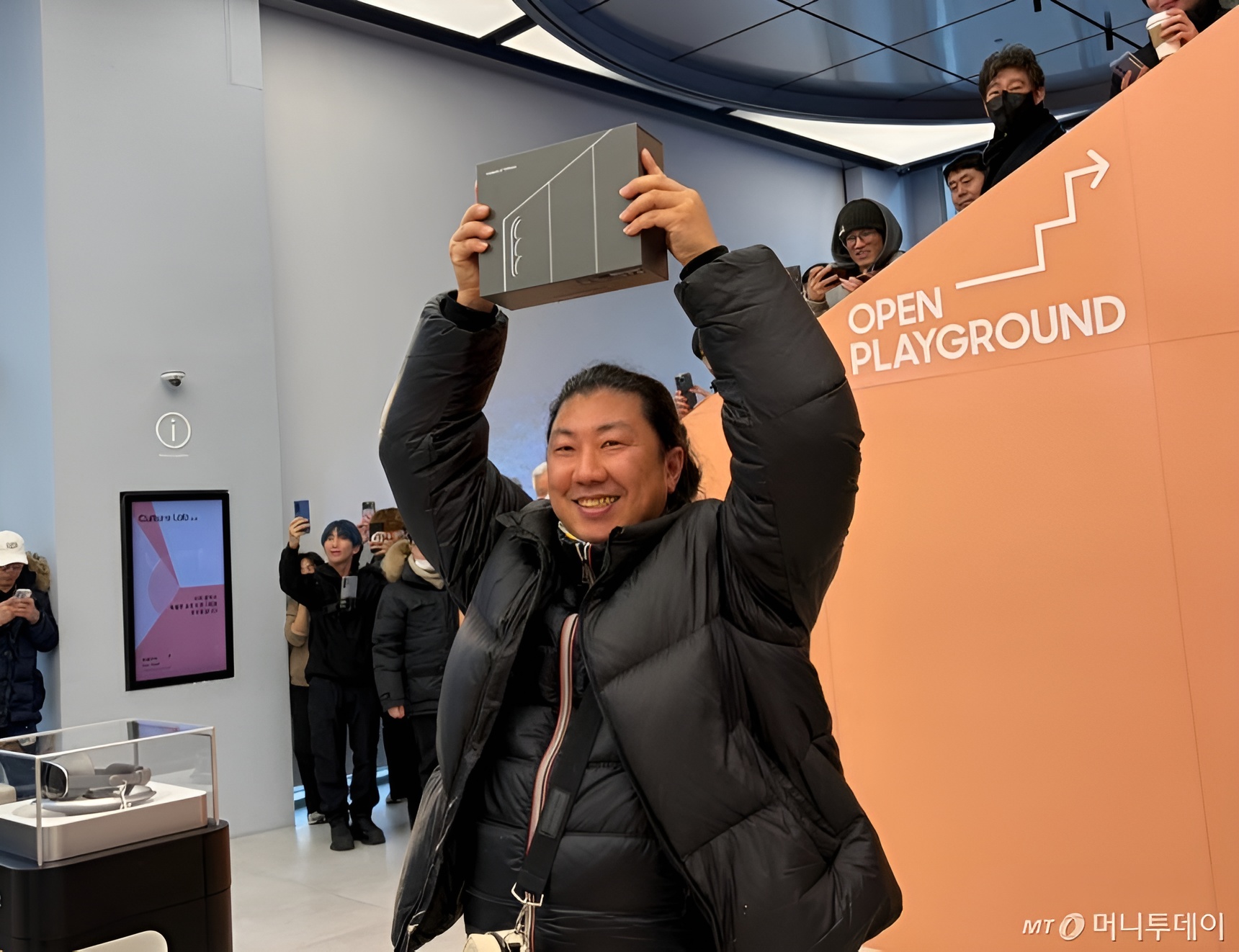A person holding a box above their head stands near a sign that says 'OPEN PLAYGROUND,' surrounded by people taking photos with smartphones.