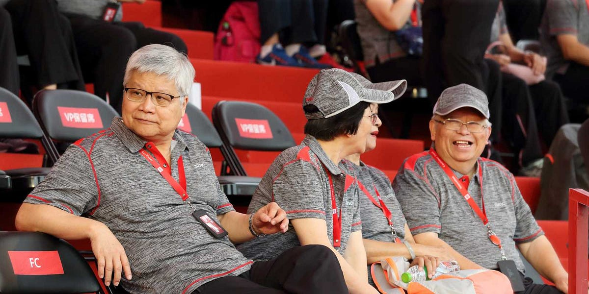 Three people are sitting on bleachers wearing matching grey shirts with red details and 'TSMC' lanyards, with empty seats labeled 'FC' and 'VIP' behind them.