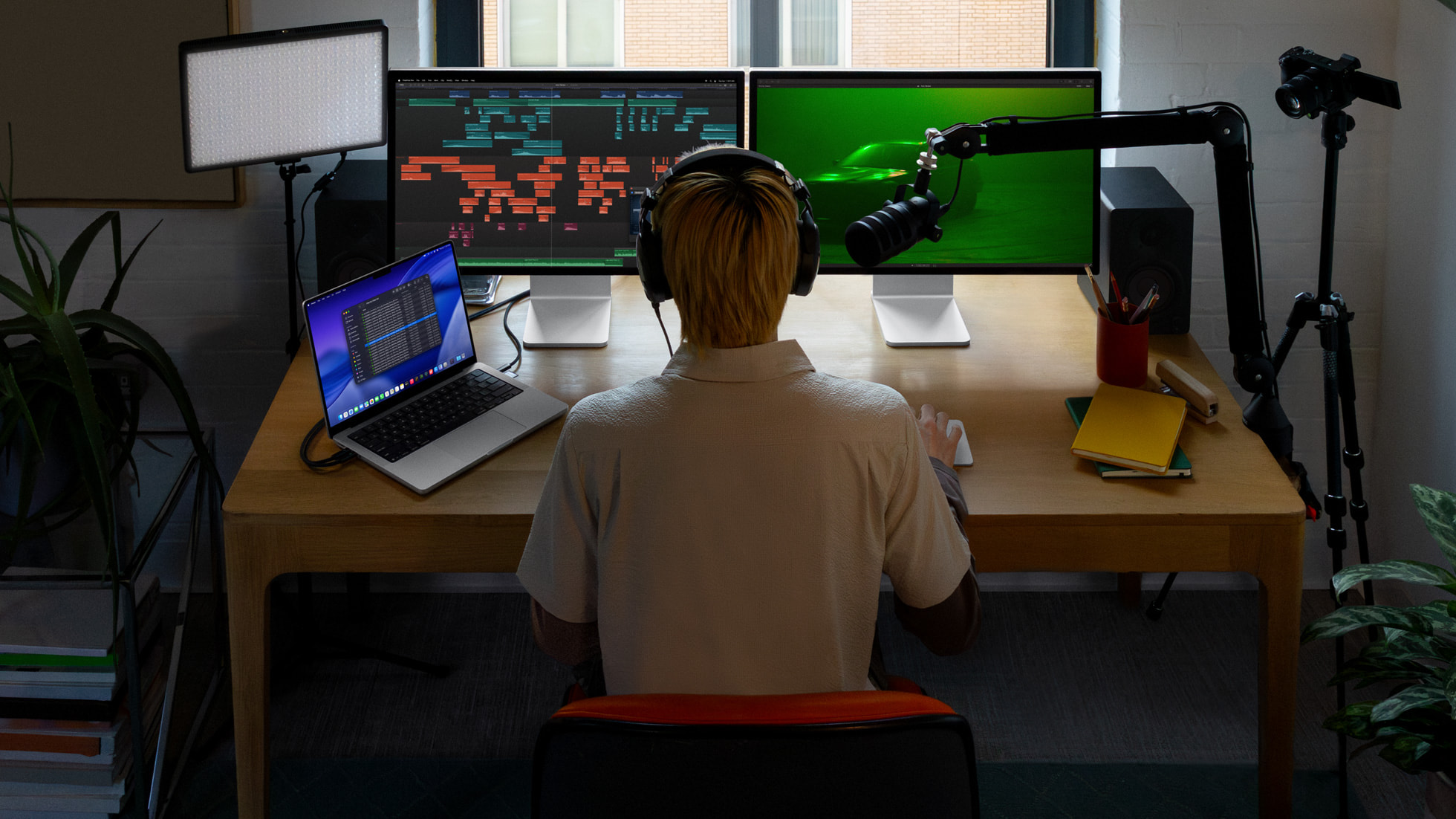 A person wearing headphones is facing dual monitors displaying a music production software, with a laptop beside them and a microphone setup on a desk.