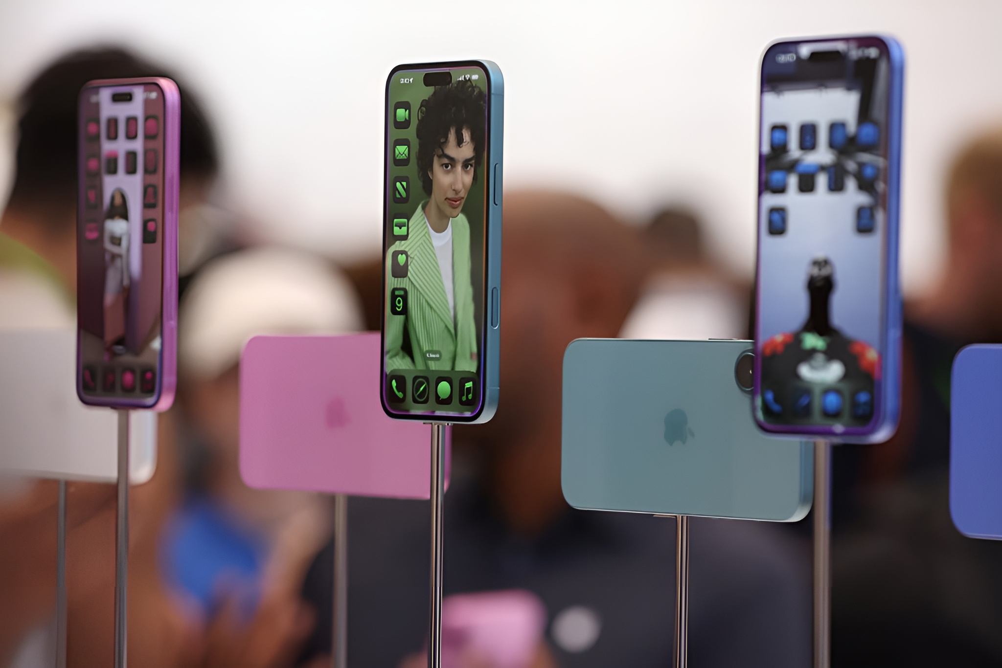 Three Apple iPhone 15 models in various colors are displayed on stands, each showing different home screens with app icons and wallpaper images.