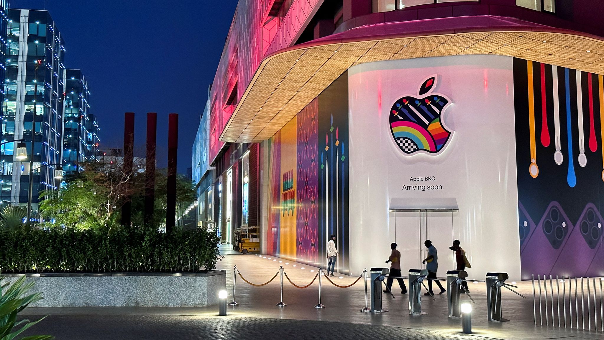 An Apple BKC store with a colorful logo on a large wall displays 'Arriving soon' at night.