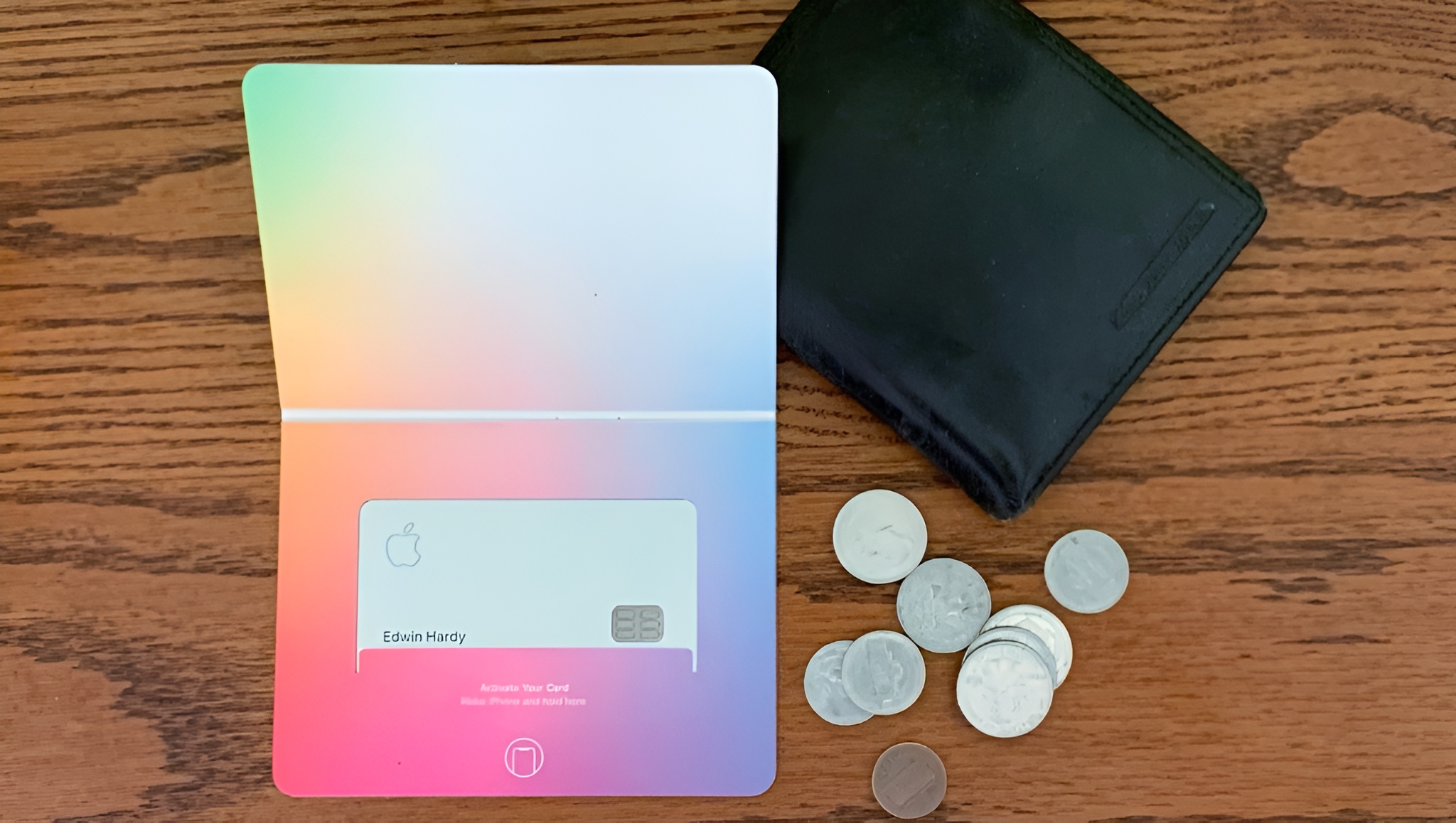 An Apple Card with the name Edwin Hardy sits in its colorful packaging next to a black wallet and several coins on a wooden surface.