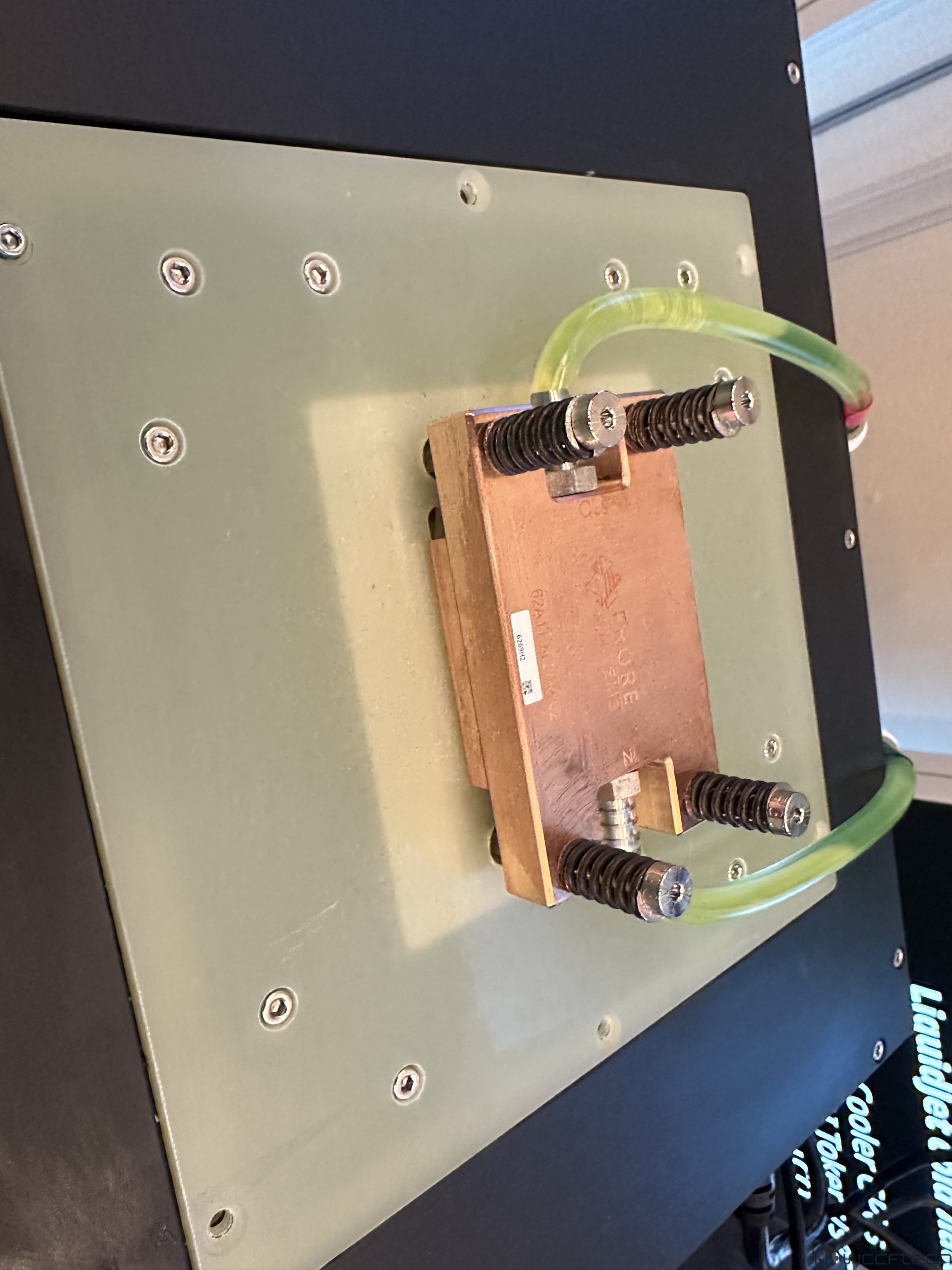 A large green circuit board is mounted with a copper coolant block connected by transparent green tubing, set against a black background displaying partially visible text about cooling.
