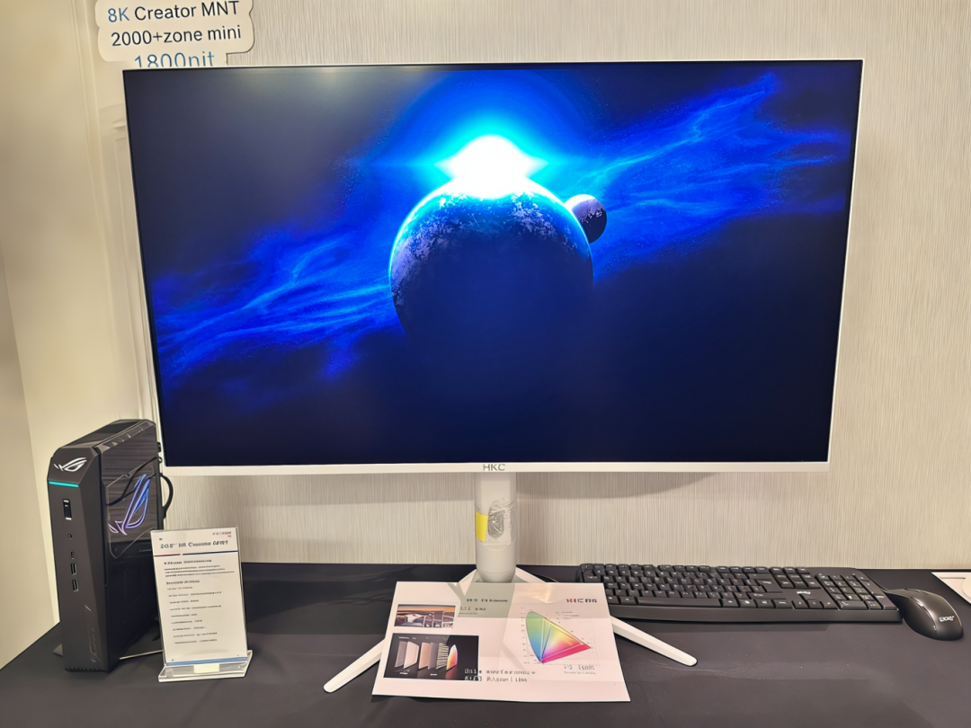 A setup with an HKC 8K Creator MNT monitor displaying a blue space scene, an ASUS ROG desktop tower, keyboard, and mouse alongside specifications and color chart paperwork on a black table.