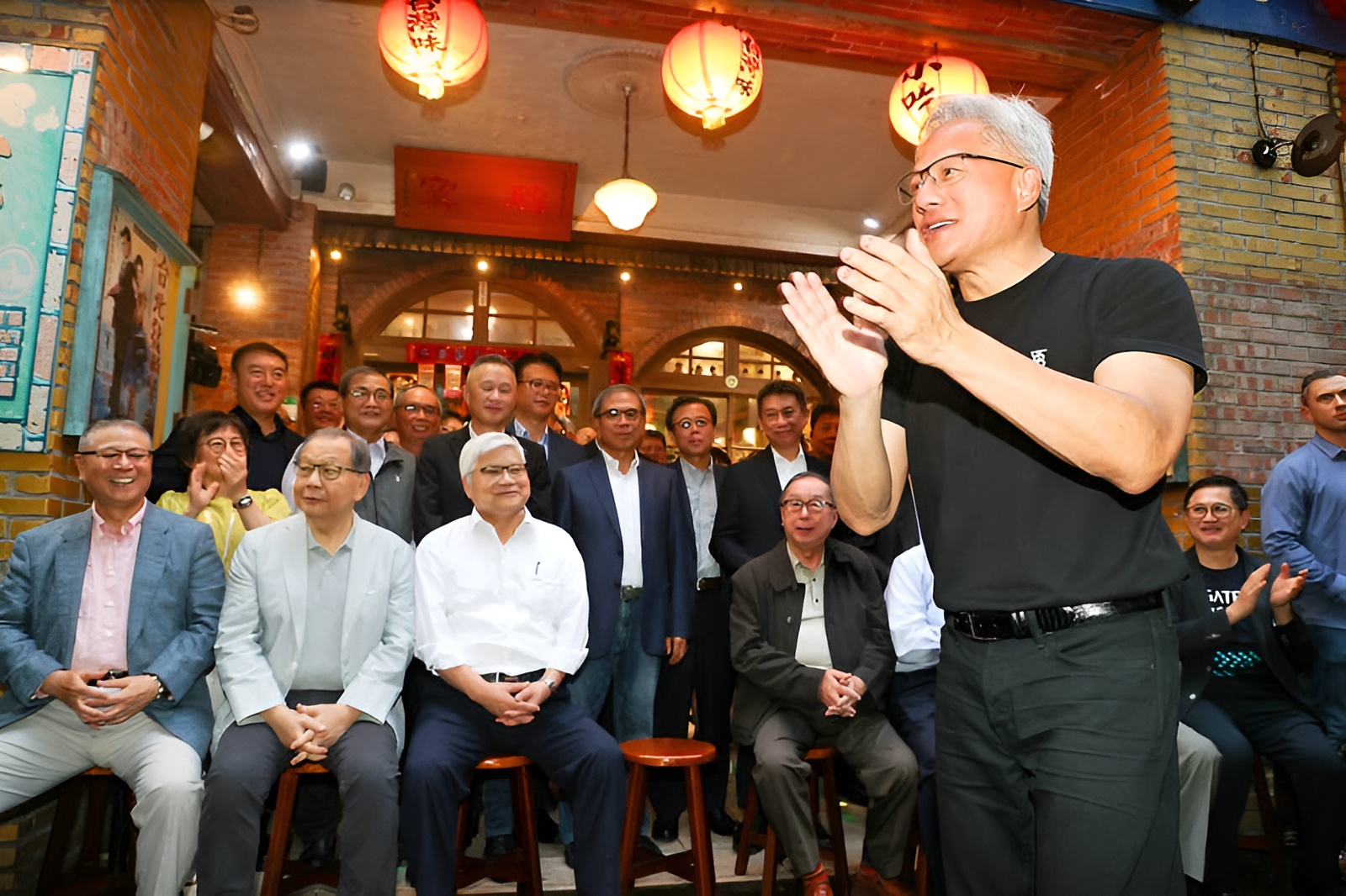 A group of people gathered indoors, with a man in a black t-shirt clapping at the front, surrounded by lanterns and a festive atmosphere.