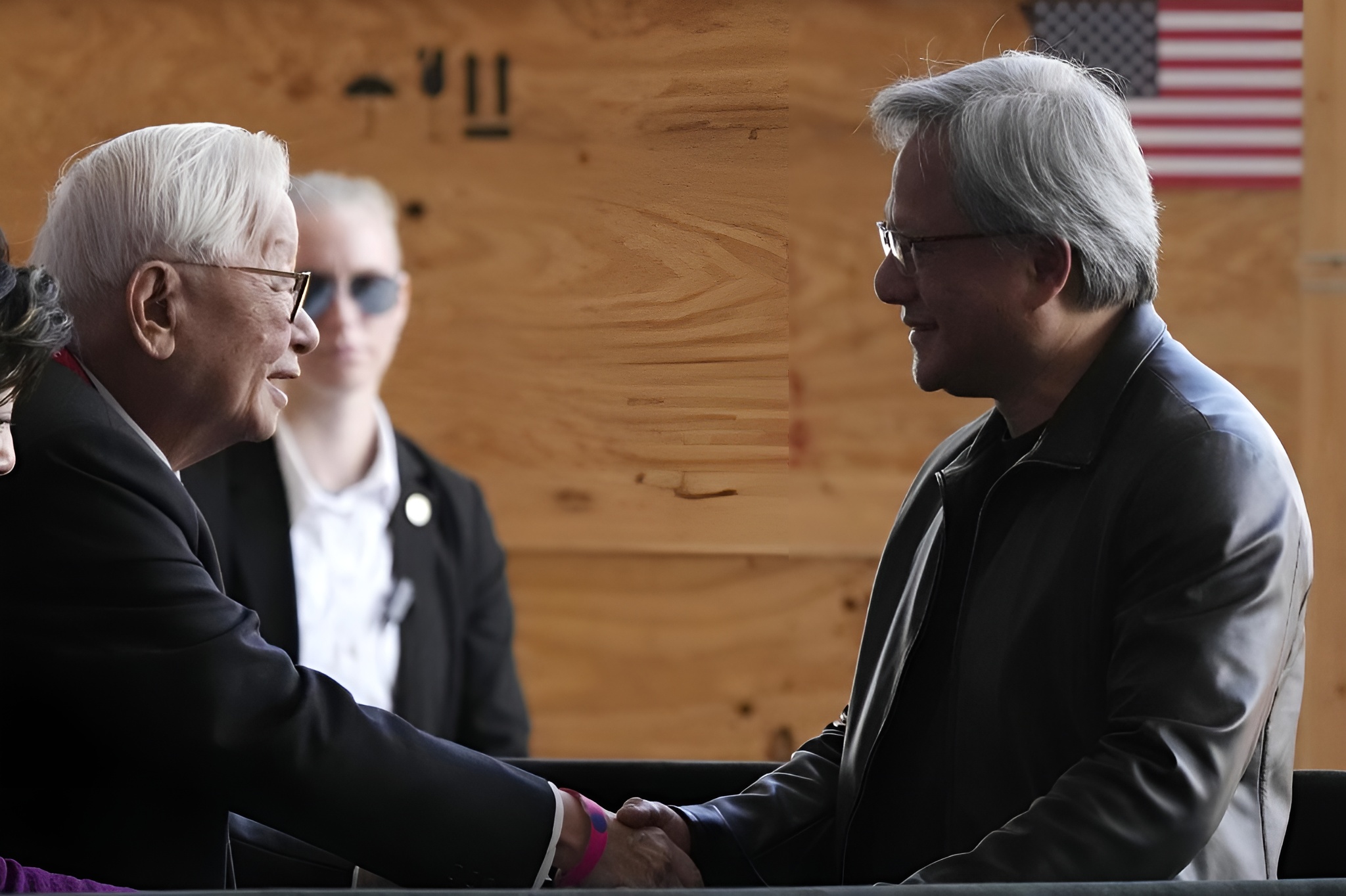 Two people, one wearing a leather jacket and glasses, shake hands in front of a wooden panel with an American flag in the