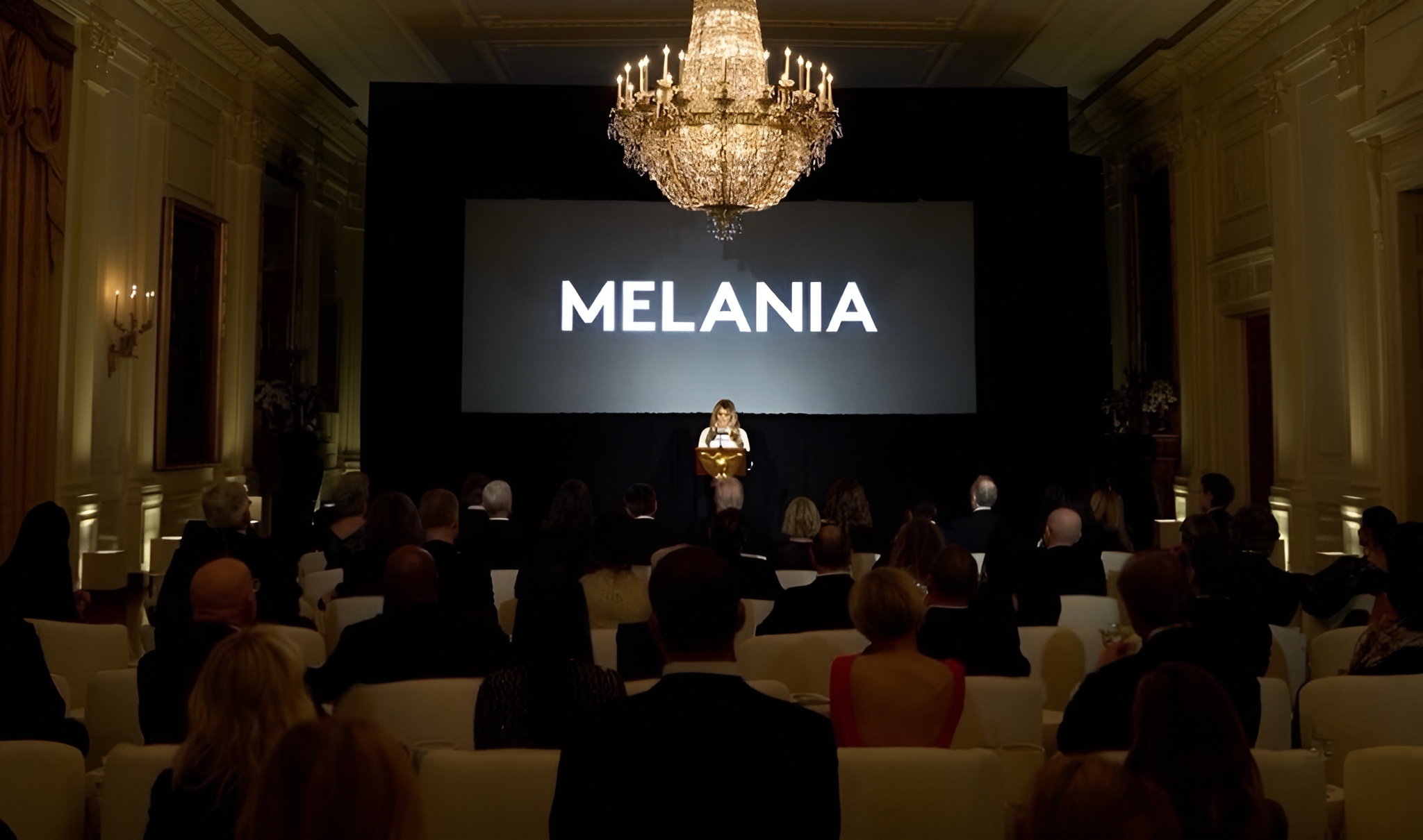 A woman stands on stage in a dimly lit room, addressing an audience with the word 'MELANIA' displayed on a large screen