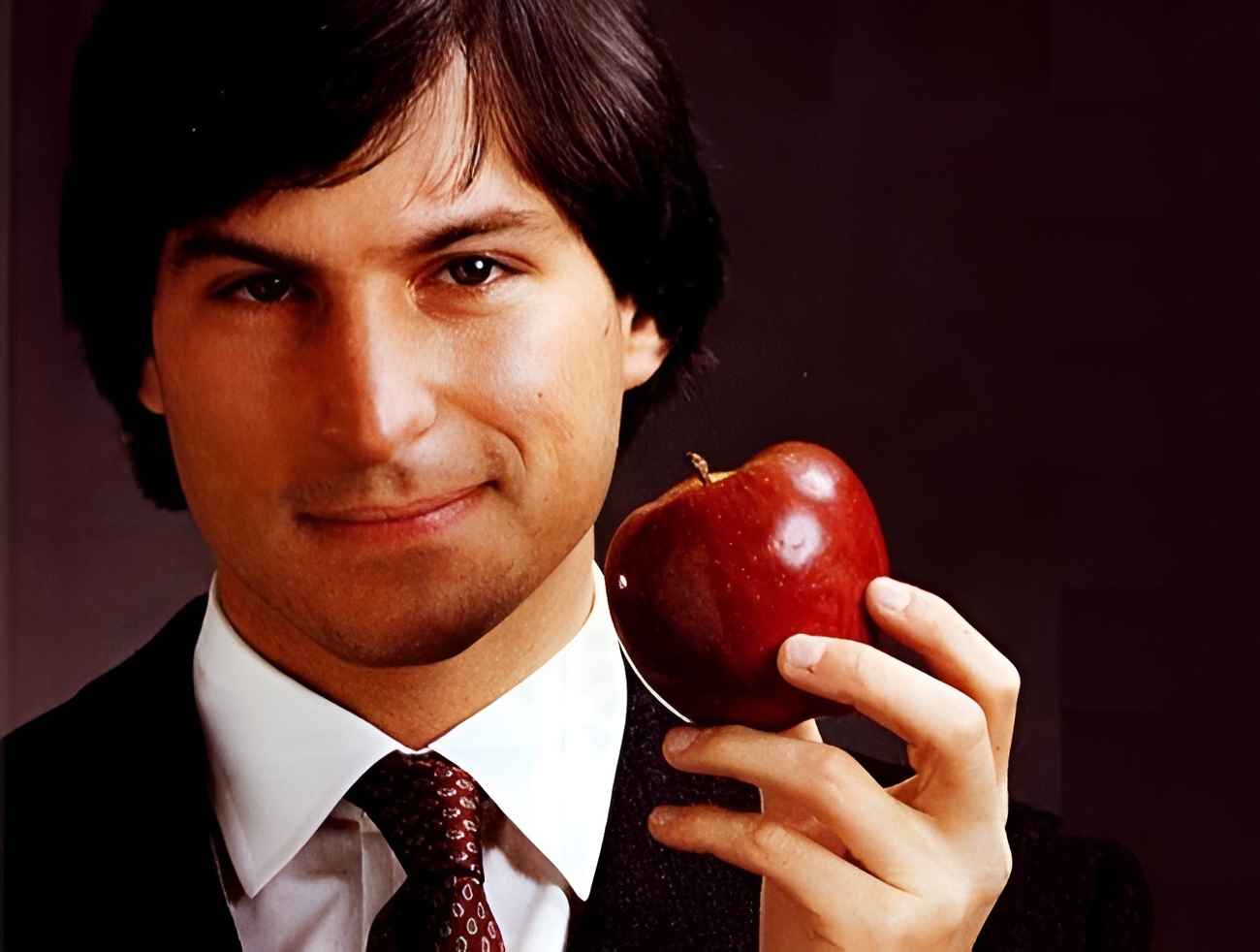 A person in a suit holding a red apple against a dark background.
