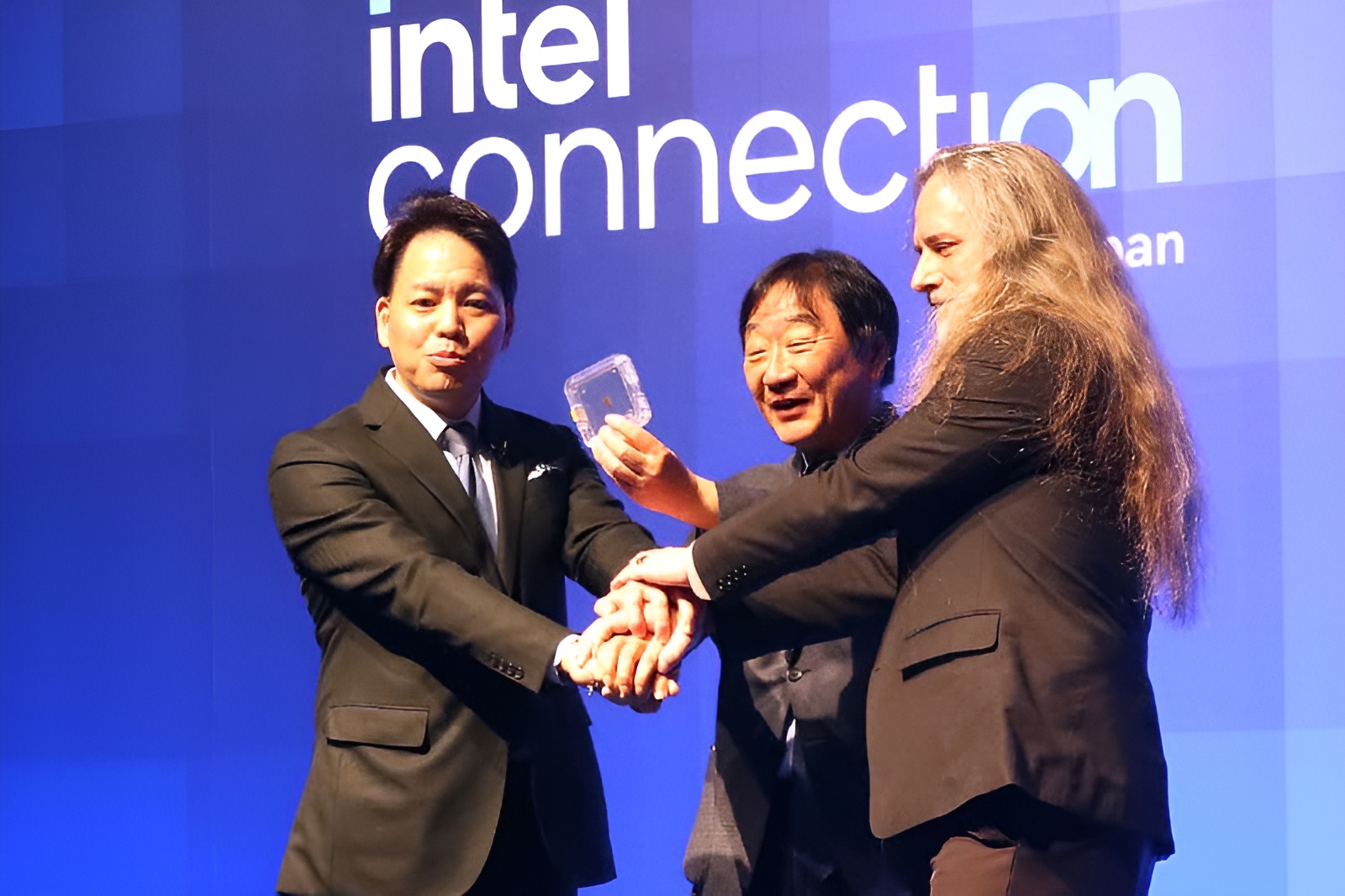 Three unidentified individuals on stage at an 'intel connection' event, with one holding a chip encased in a clear square