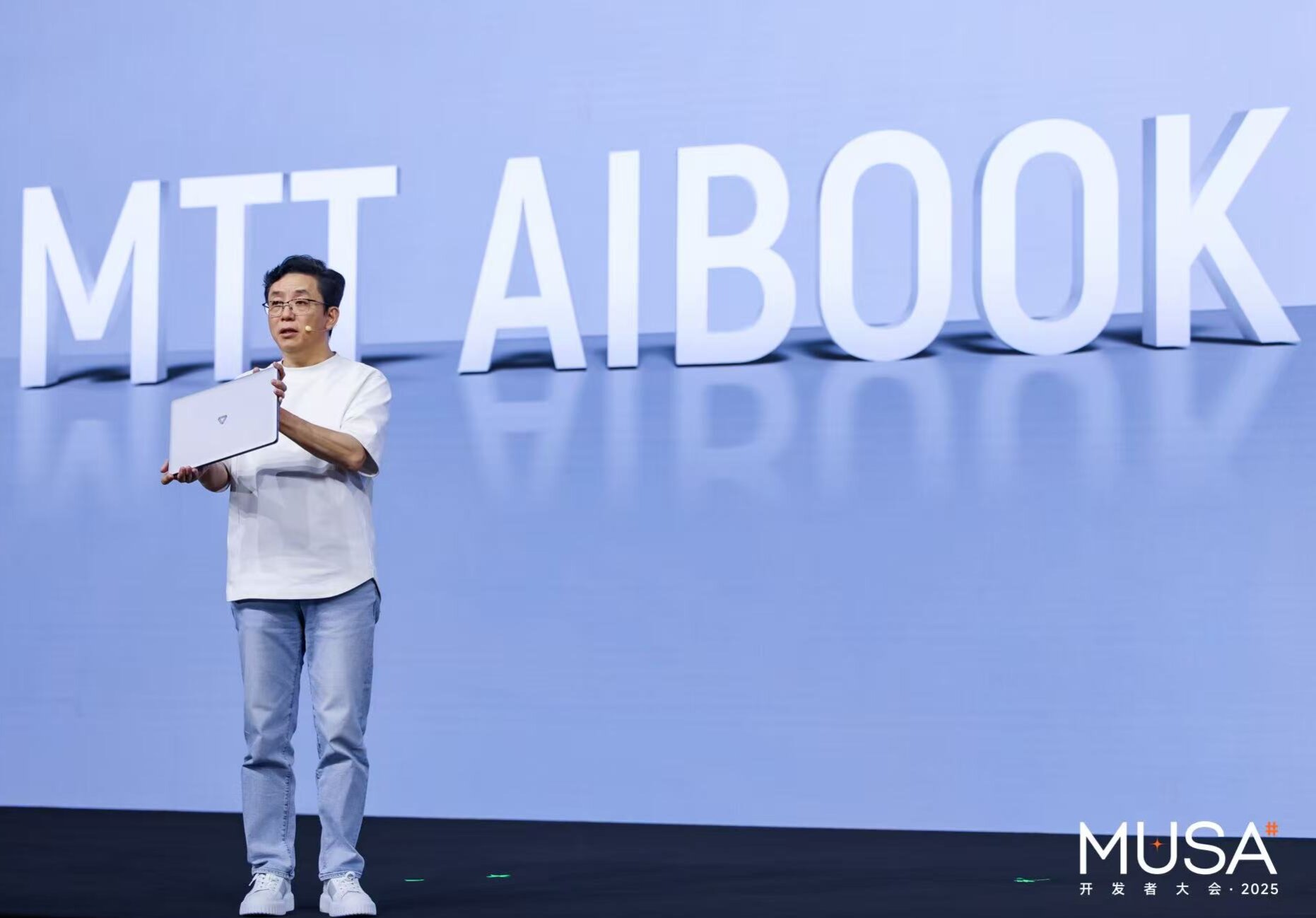 A person holding a white laptop stands on stage with large text in the background saying 'MTT Aibook' and a conference logo
