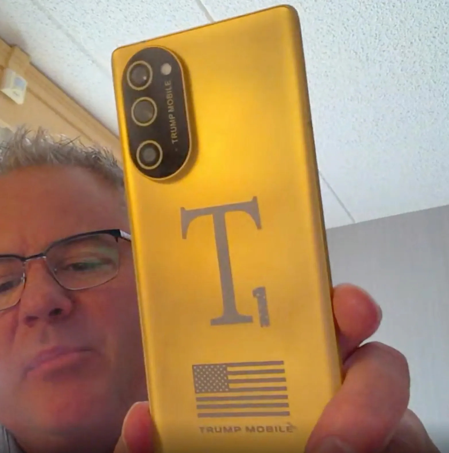 A person holding a gold Trump Mobile phone with a large 'T1' logo and an American flag on its back.