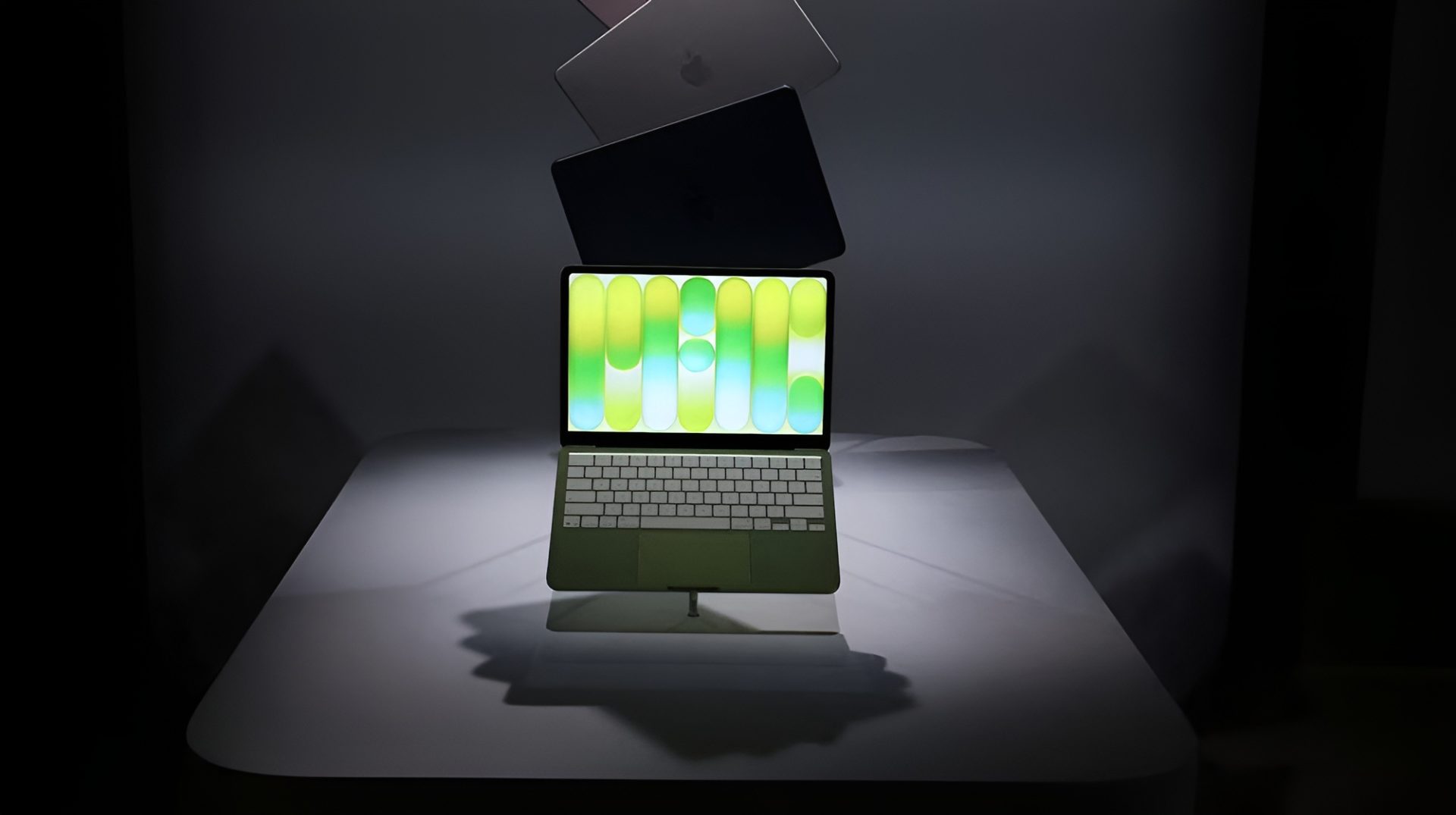 A green Apple MacBook with a colorful display is prominently showcased in front of several other stacked laptops.