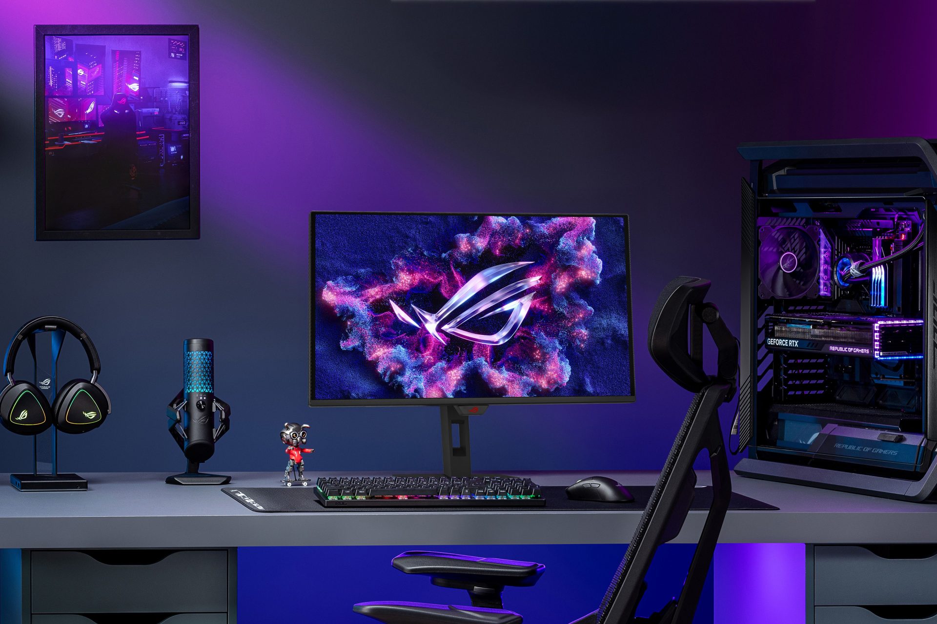 A gaming setup featuring an ASUS ROG monitor displaying the ROG logo, a GeForce RTX graphics card inside a desktop PC, ROG headphones on a stand, and a ROG-themed microphone on a desk.