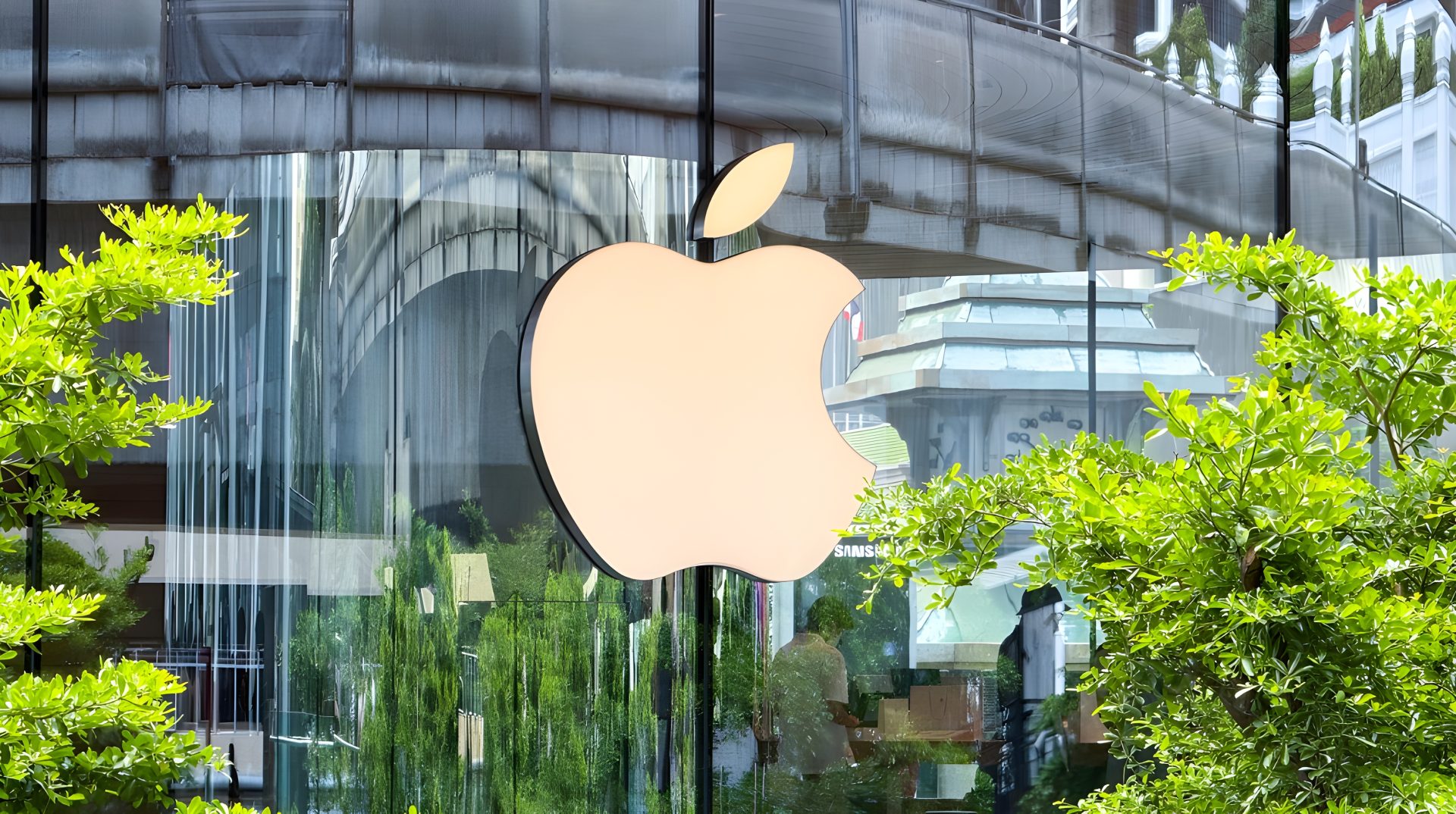 A large Apple logo is displayed on the glass facade of an Apple Store surrounded by greenery and reflecting nearby buildings.