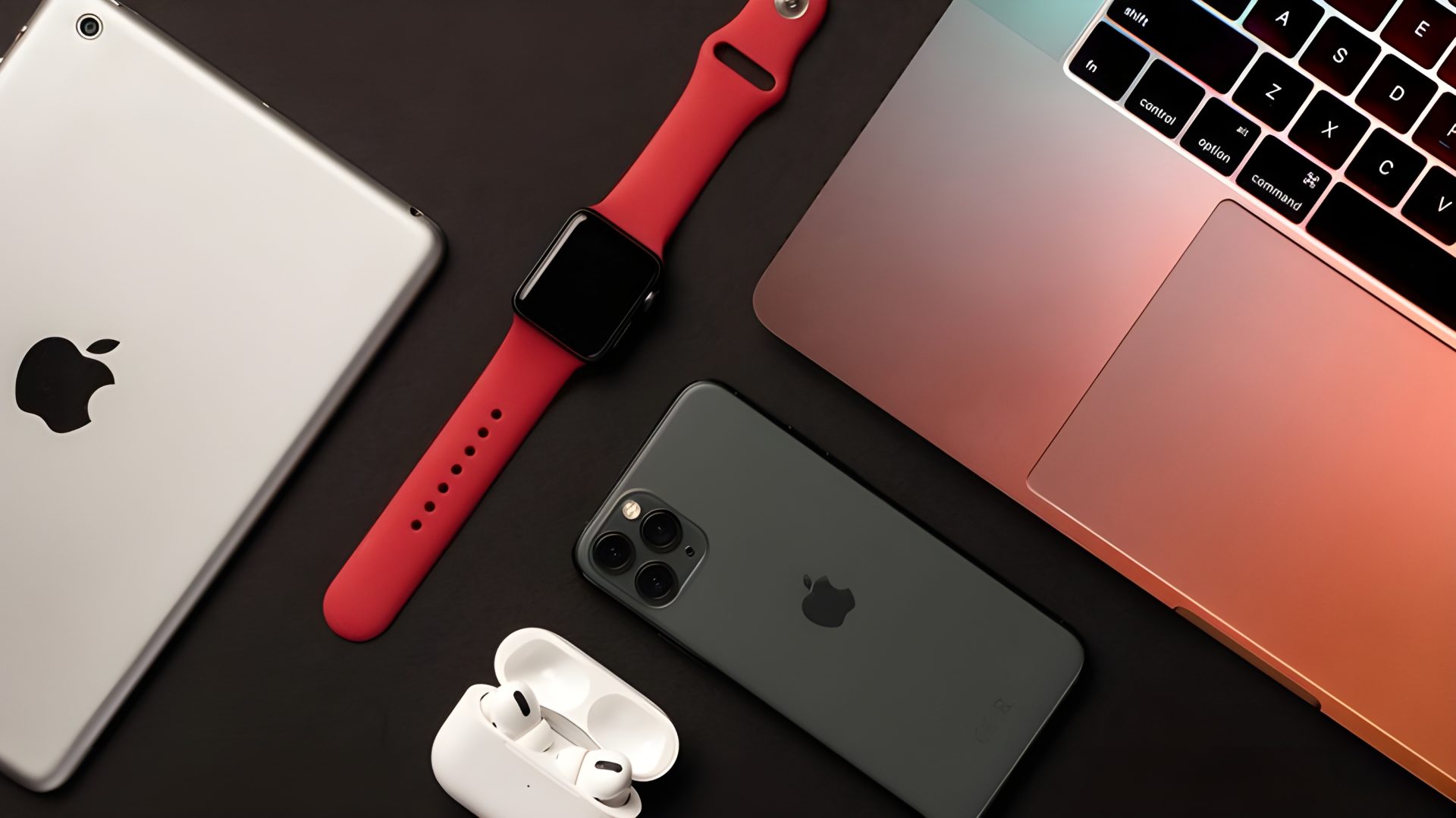 An Apple iPad, Apple Watch with a red band, iPhone, AirPods, and MacBook on a dark surface.