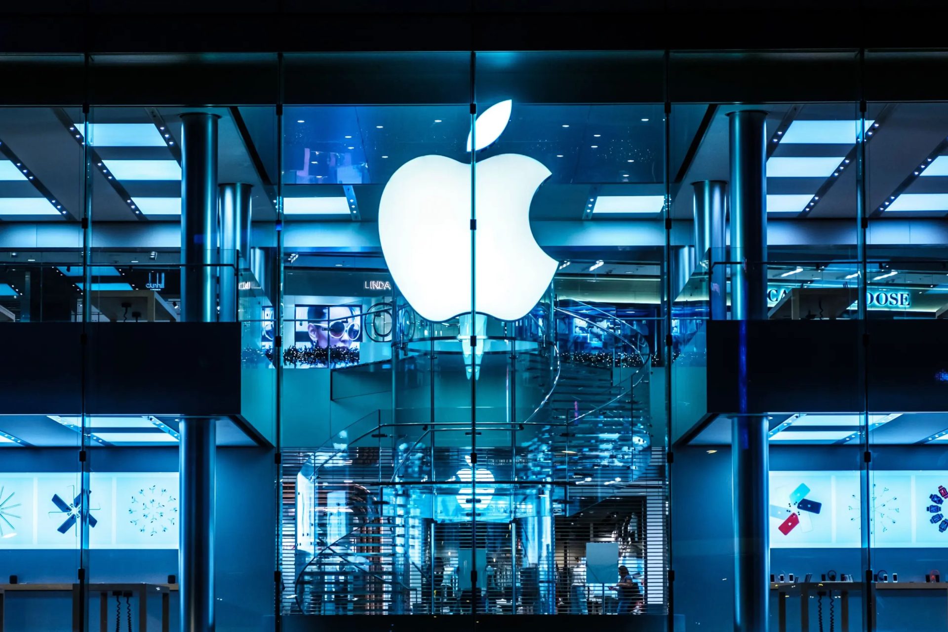 The exterior of an Apple Store displays a large illuminated Apple logo against a modern, glass-fronted building.