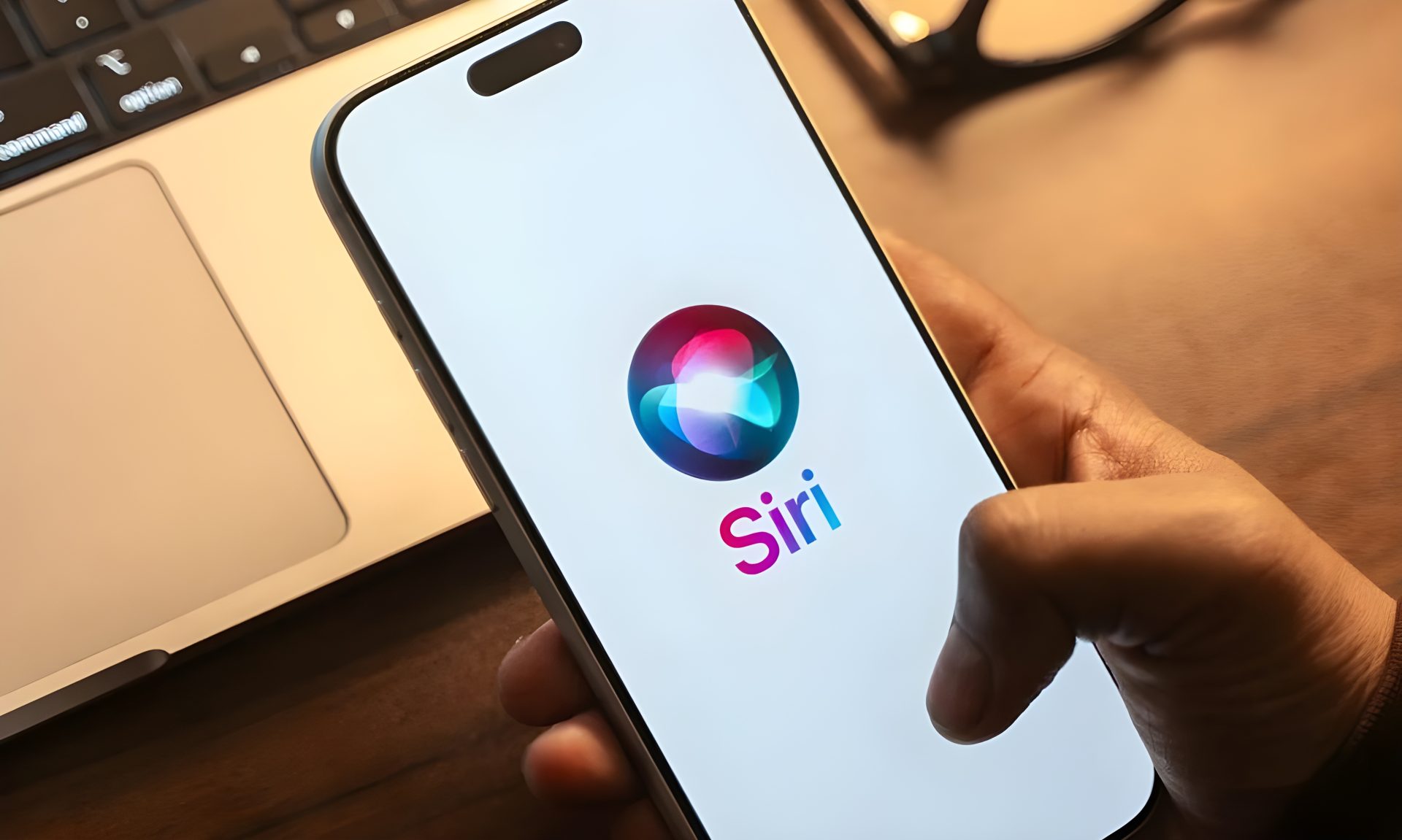 A person holding a smartphone displaying the Siri logo and text, with a laptop visible in the background.