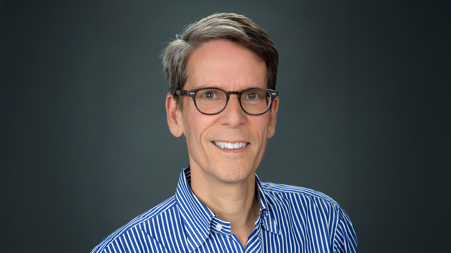 A person wearing glasses and a blue striped shirt is smiling against a dark background.