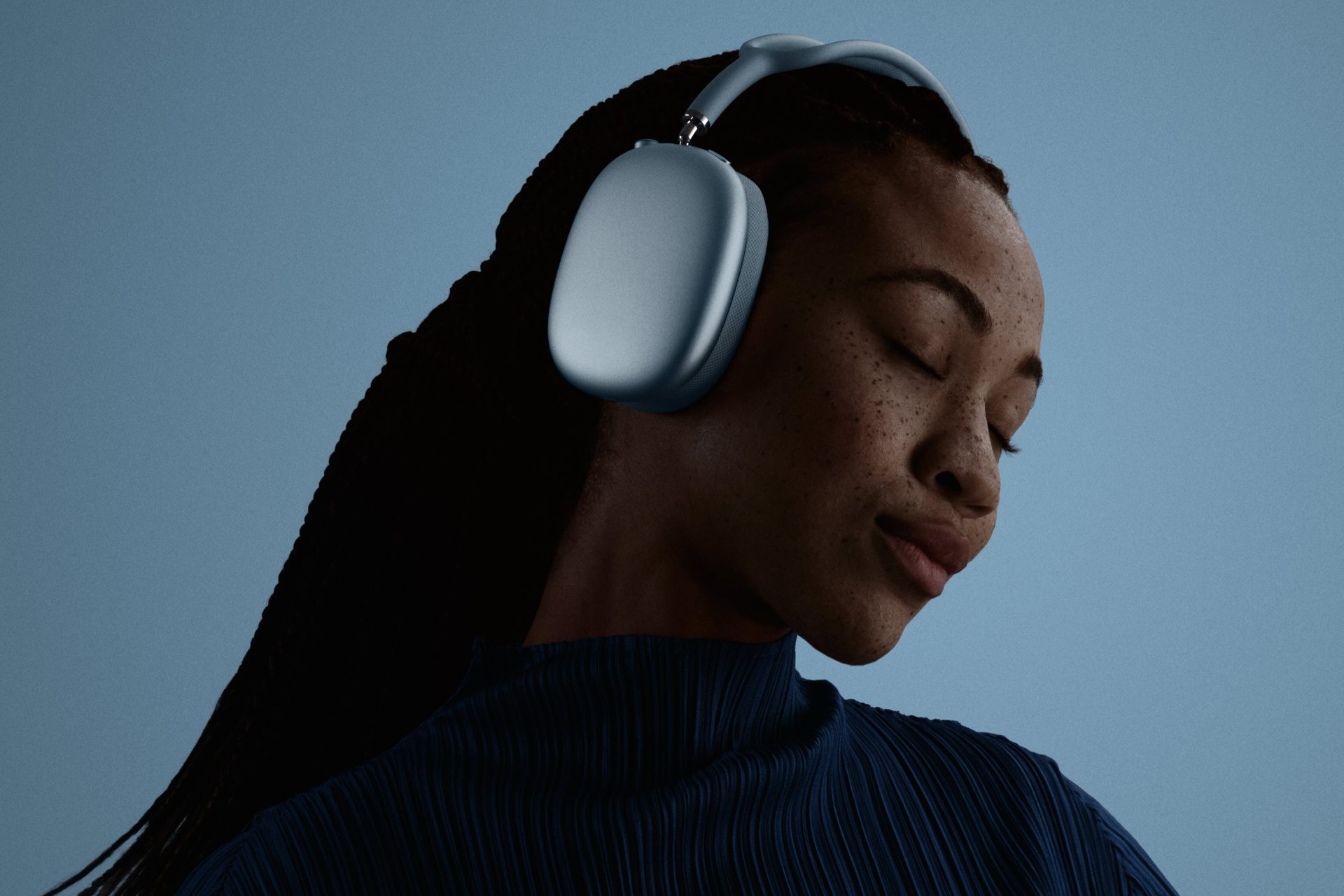 A person wearing Apple AirPods Max headphones with eyes closed, set against a plain blue background.