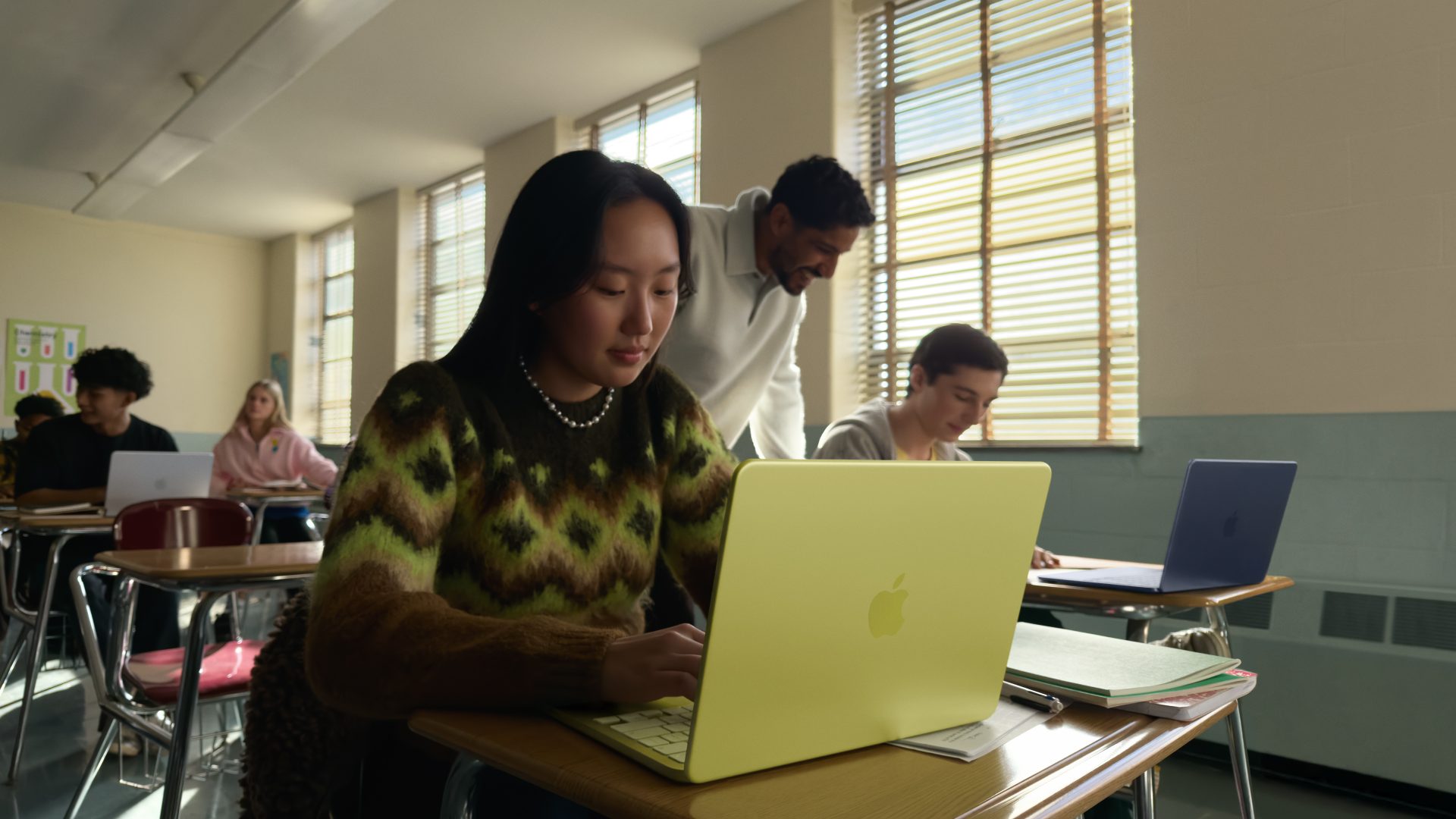 A classroom setting with students using Apple MacBook laptops in pastel colors, including a yellow one in the foreground.