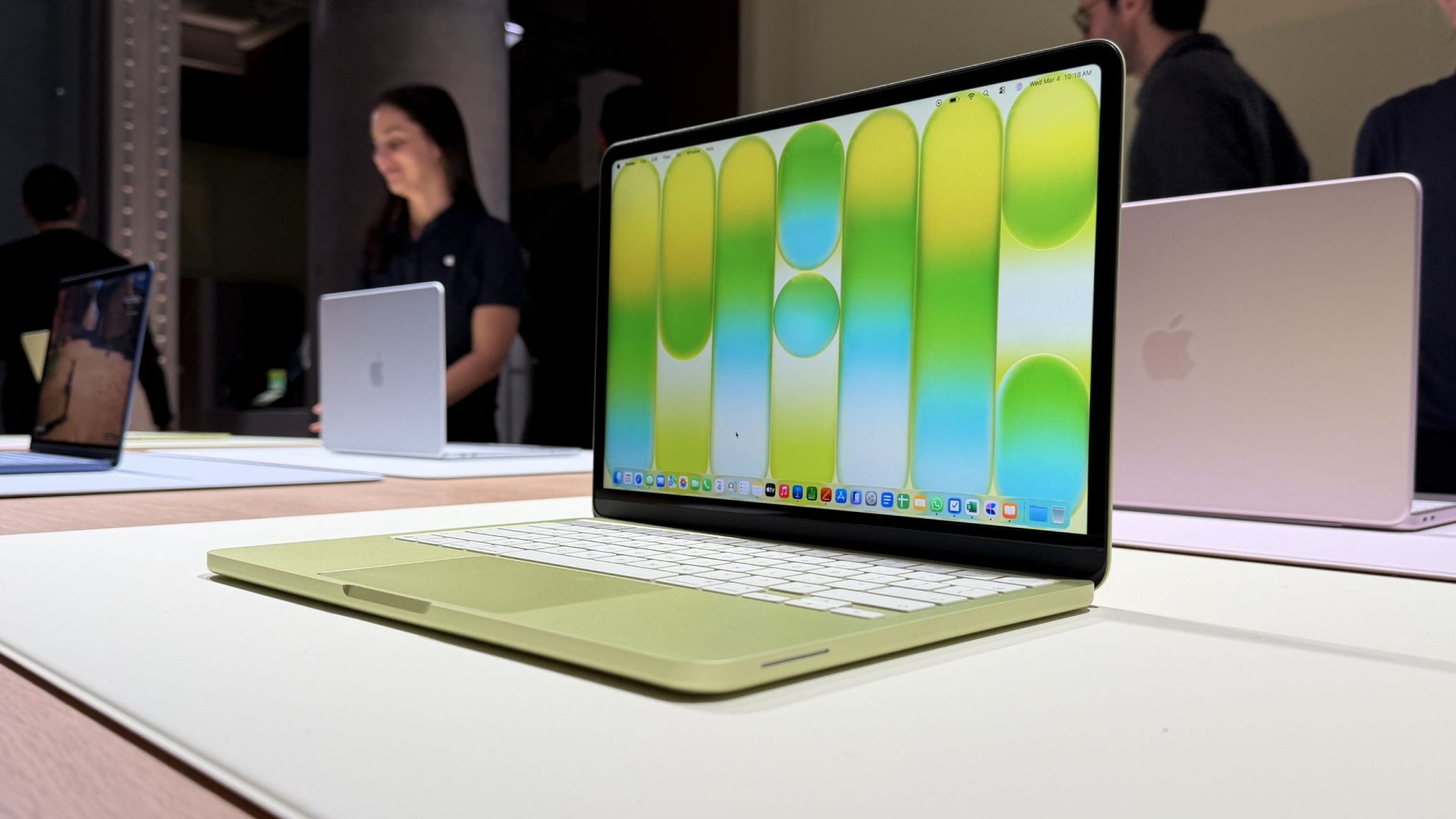 A green Apple MacBook Air with a colorful abstract desktop wallpaper displayed on a table, surrounded by other similar laptops and a blurred background of people.