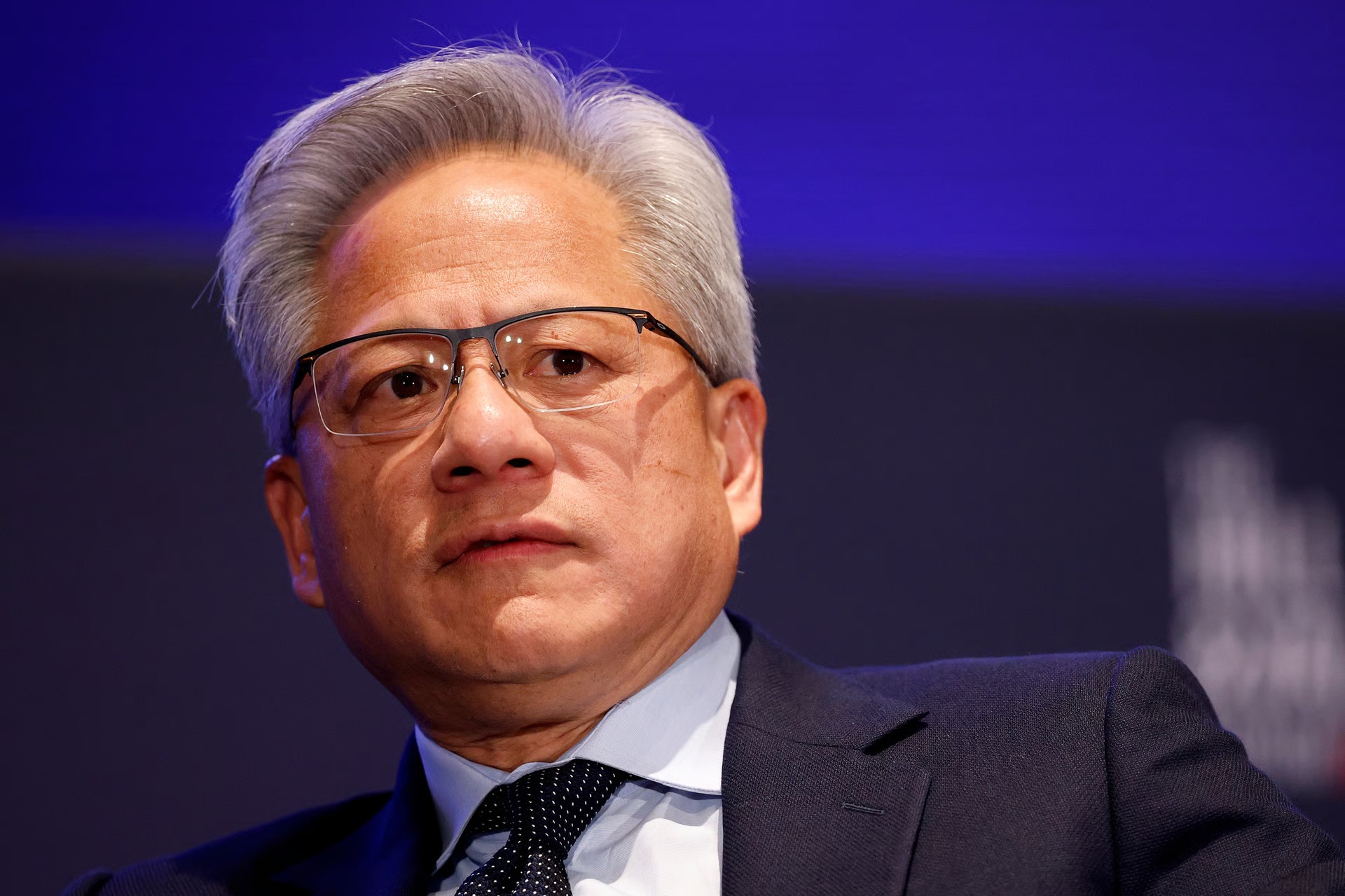 A man in glasses and a suit is looking towards the right against a dark blue background.