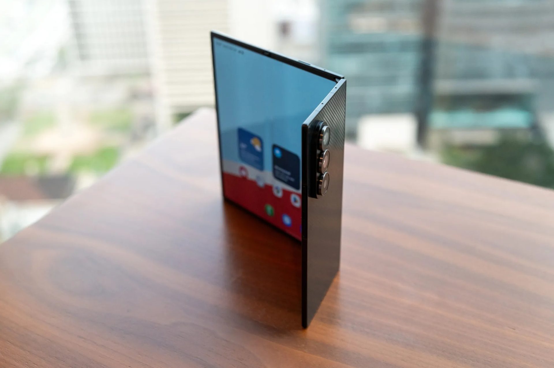 A foldable smartphone standing upright showing its hinge and rear camera setup with an app interface visible on the partially unfolded display.