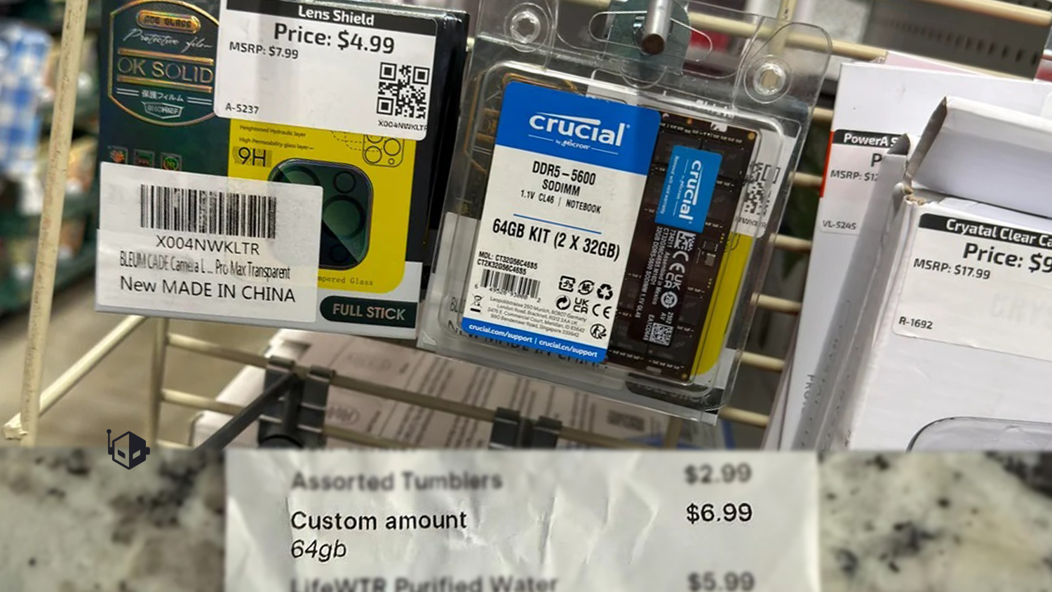 A display rack shows a Crucial DDR5-5600 SODIMM 64GB Kit (2 x 32GB) alongside various product packaging, with visible prices like '$4.99' and '$9.99.'