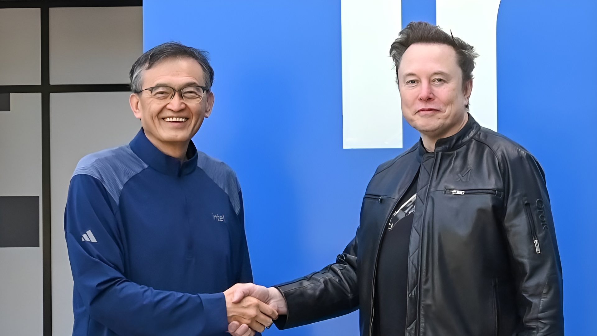 Two men shaking hands, with one wearing an Intel branded jacket, standing in front of a blue background.