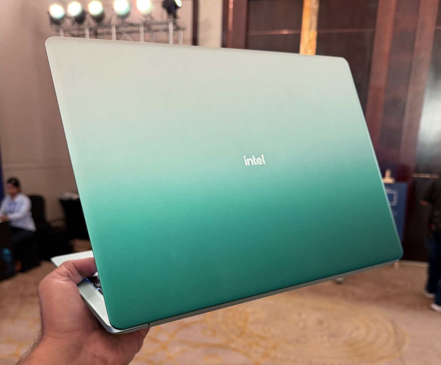 A hand holds a green-gradient laptop with the 'intel' logo on its lid in a conference setting.