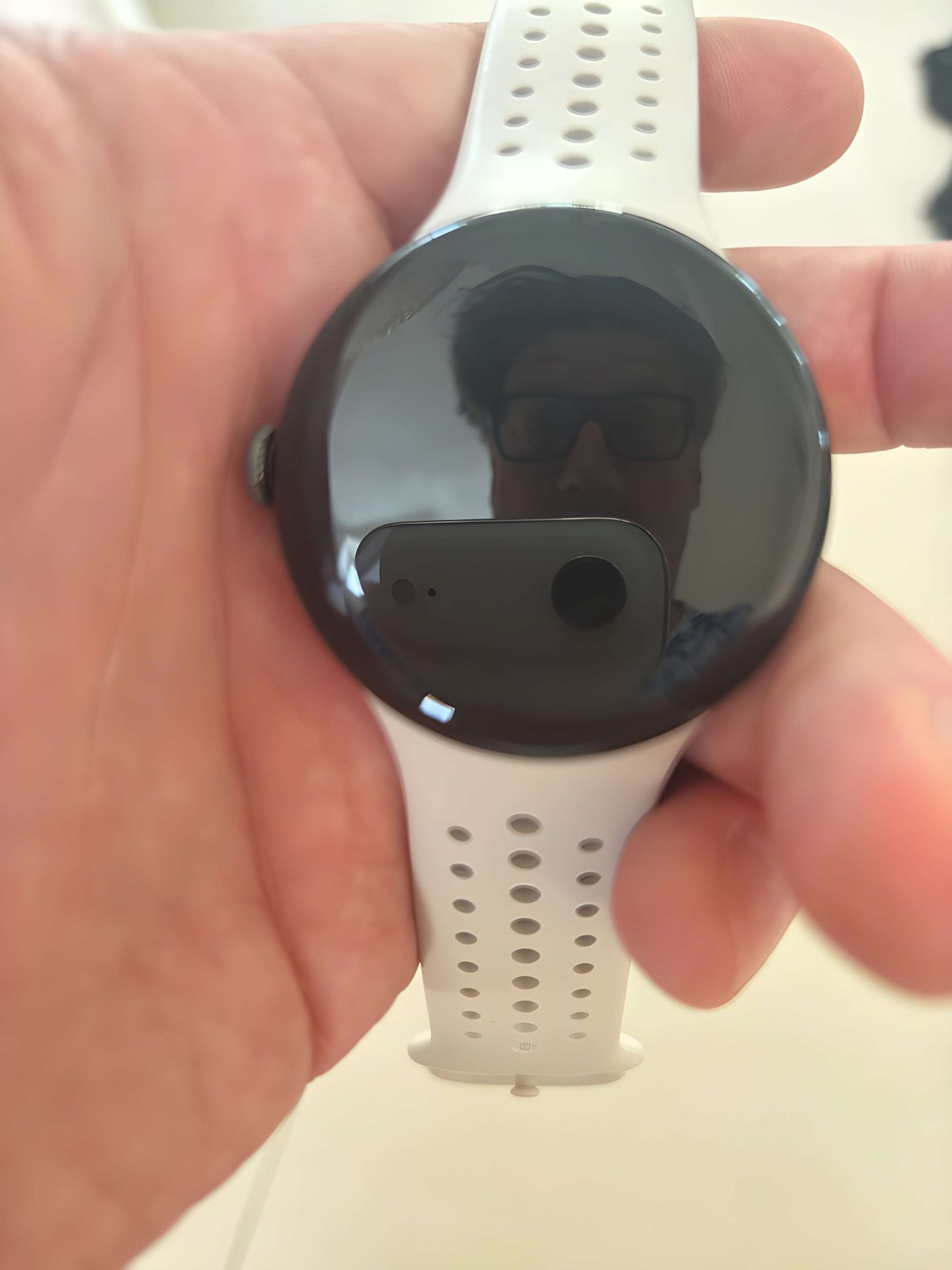 A person holds a Google Pixel Watch with a white perforated strap, visible through a reflection on the watch face.