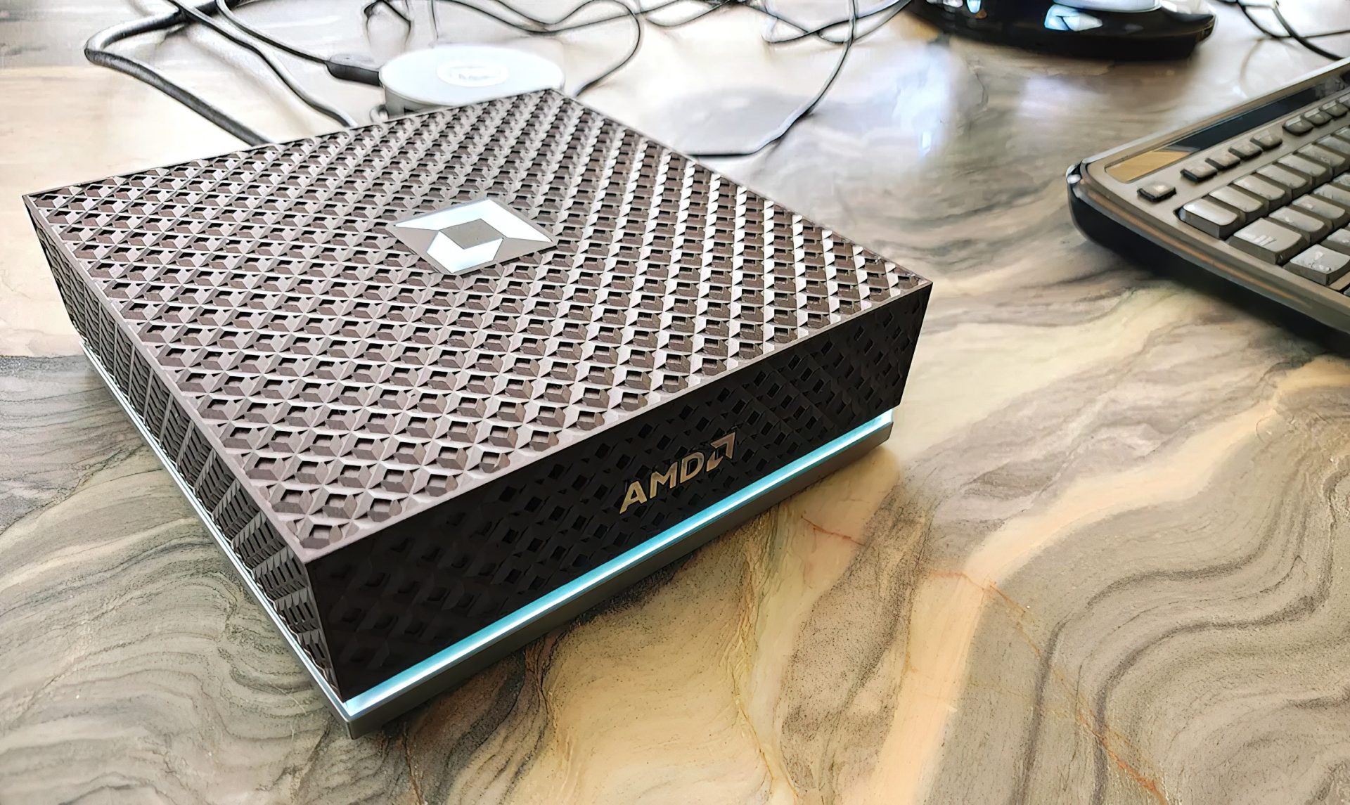 A compact device with a textured surface featuring the AMD logo on the front, placed on a marble table next to a keyboard.