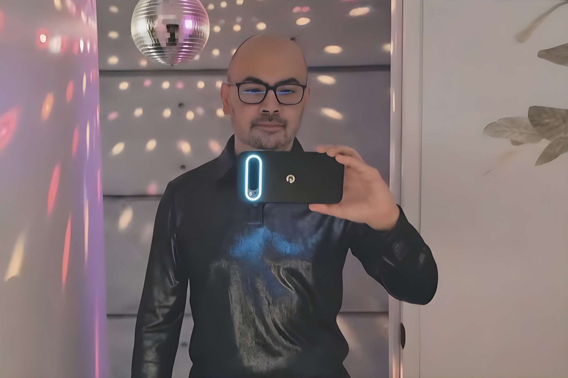A person wearing a black shirt takes a selfie in a room with a disco ball and colorful light reflections, holding a smartphone with a glowing camera module.