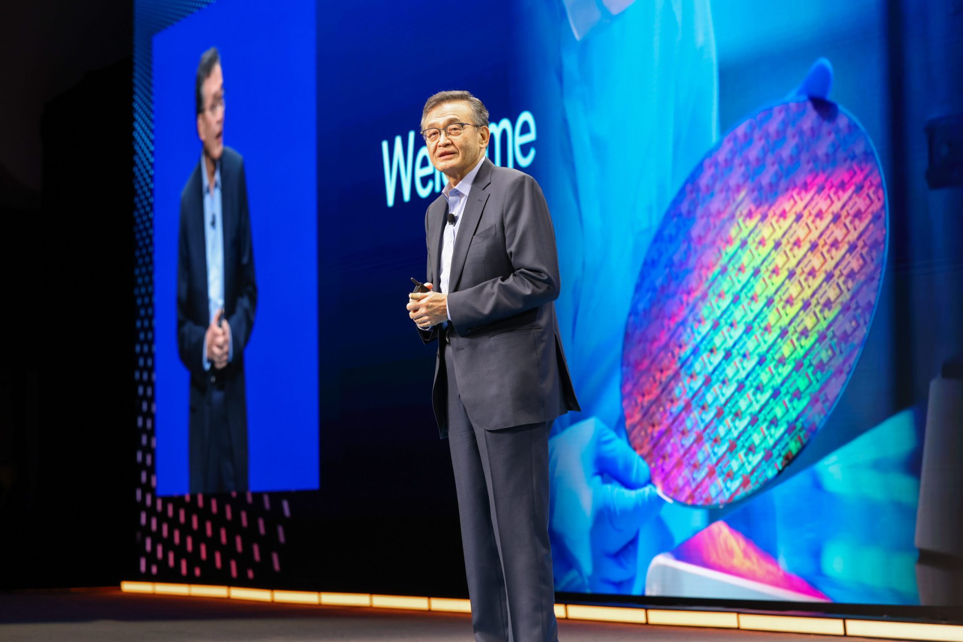 A person in a suit stands on stage with a large semiconductor wafer image displayed behind them, and the word 'Welcome' visible on a screen.