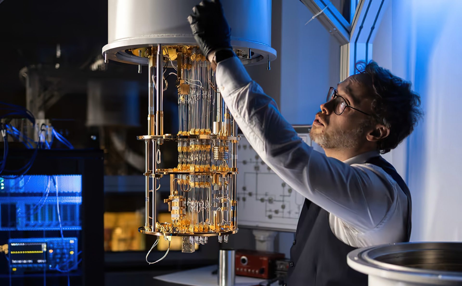 A person in a lab adjusts a complex quantum computing device suspended from above in a dimly lit laboratory setting.