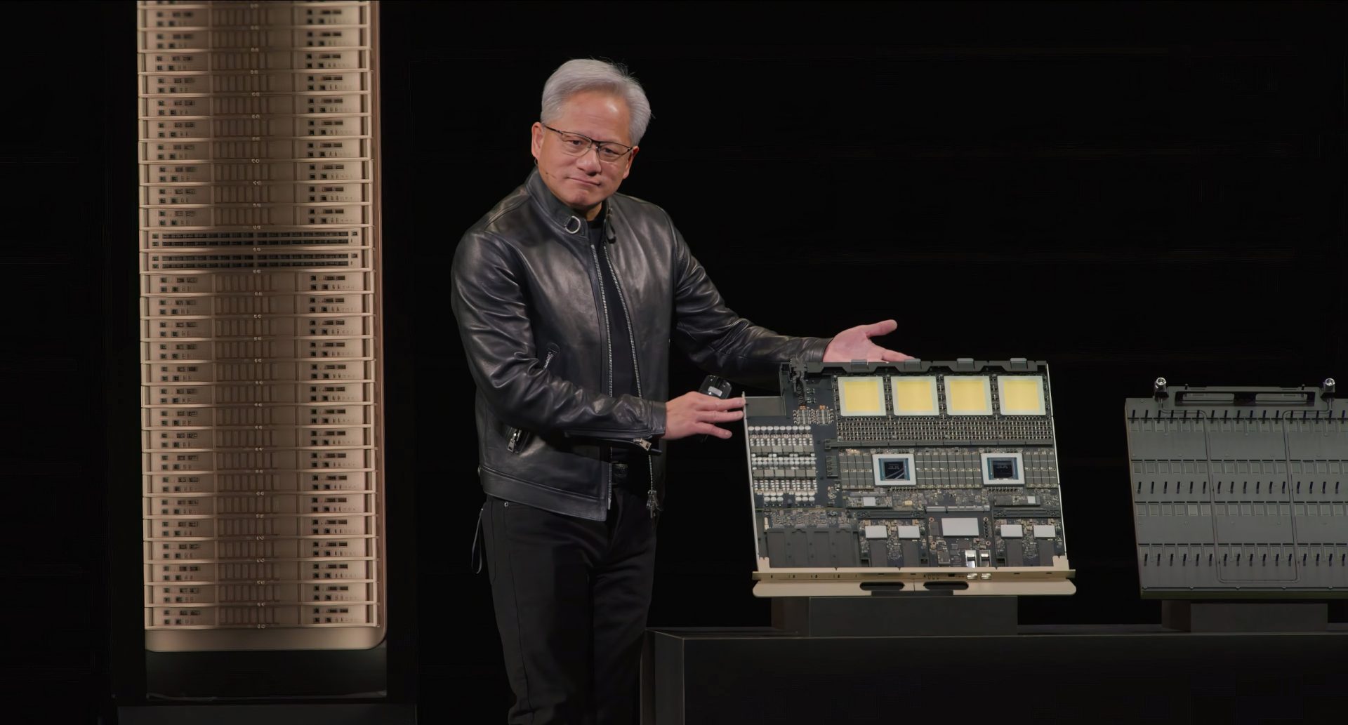 An individual in a leather jacket is presenting a large circuit board design next to a stack of horizontal units.