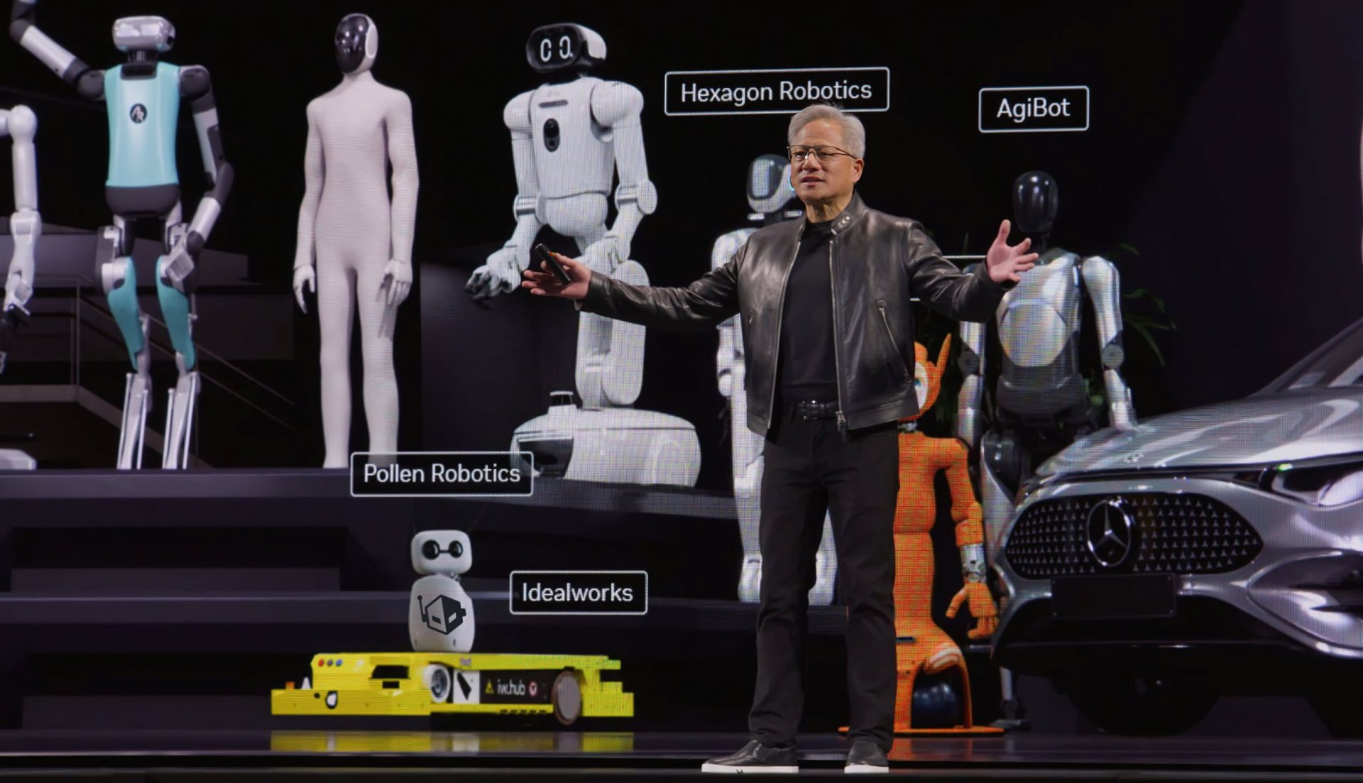 A presenter in a black jacket stands on stage illustrated with several robotic models labeled 'Hexagon Robotics,' 'AgiBot,' 'Pollen Robotics,' and 'Idealworks,' alongside a futuristic vehicle front.