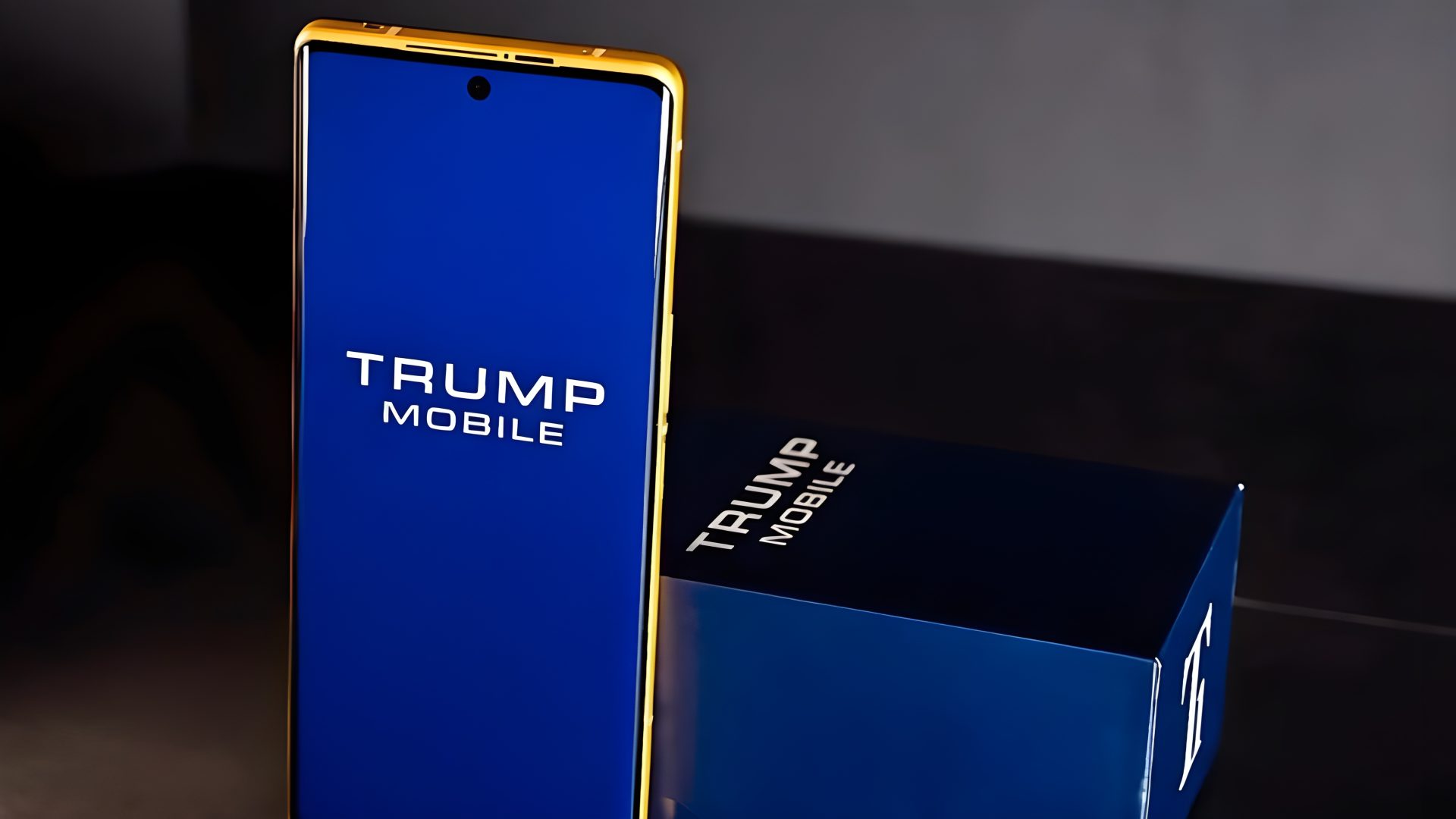 A gold-trimmed smartphone with 'TRUMP MOBILE' displayed on its blue screen stands next to a matching box.